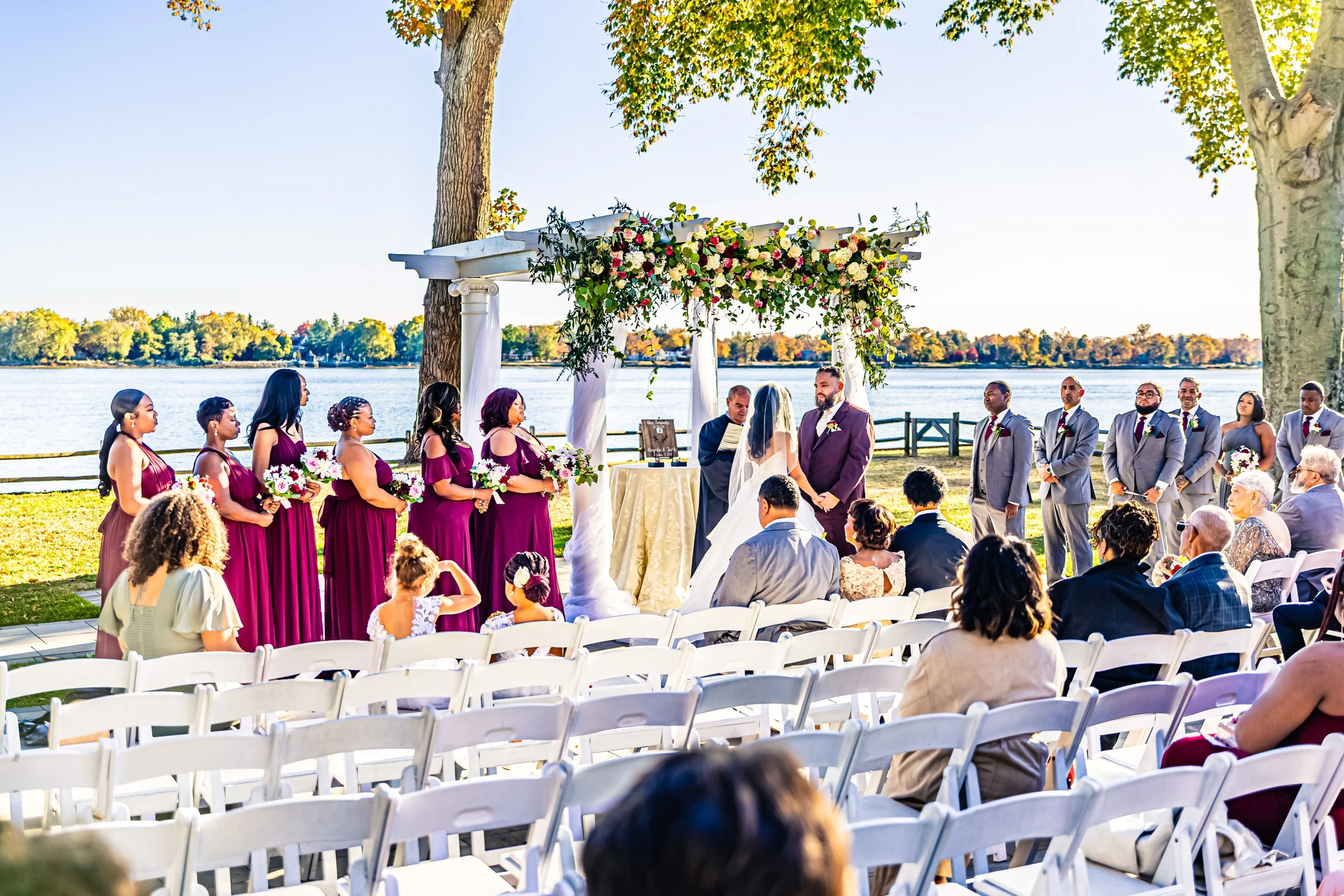 A wedding ceremony taking place outdoors by a lake with a white floral arch, officiant, bride in a white gown, groom in a purple suit, wedding party in purple and gray suits, guests seated in white chairs, large trees, and a clear blue sky.