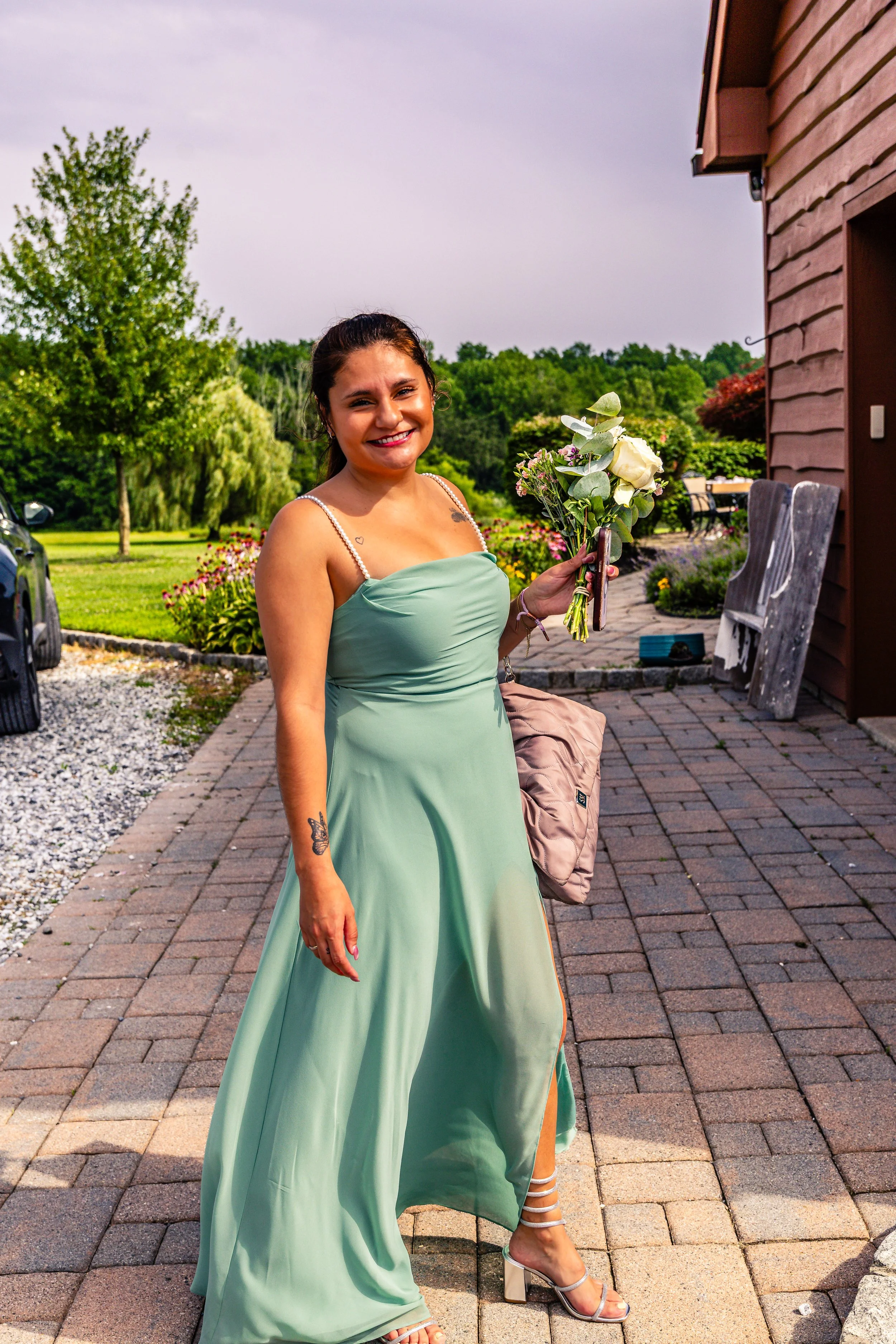A woman in a light green dress holding a bouquet of white and pink flowers, smiling outdoors on a sunny day.