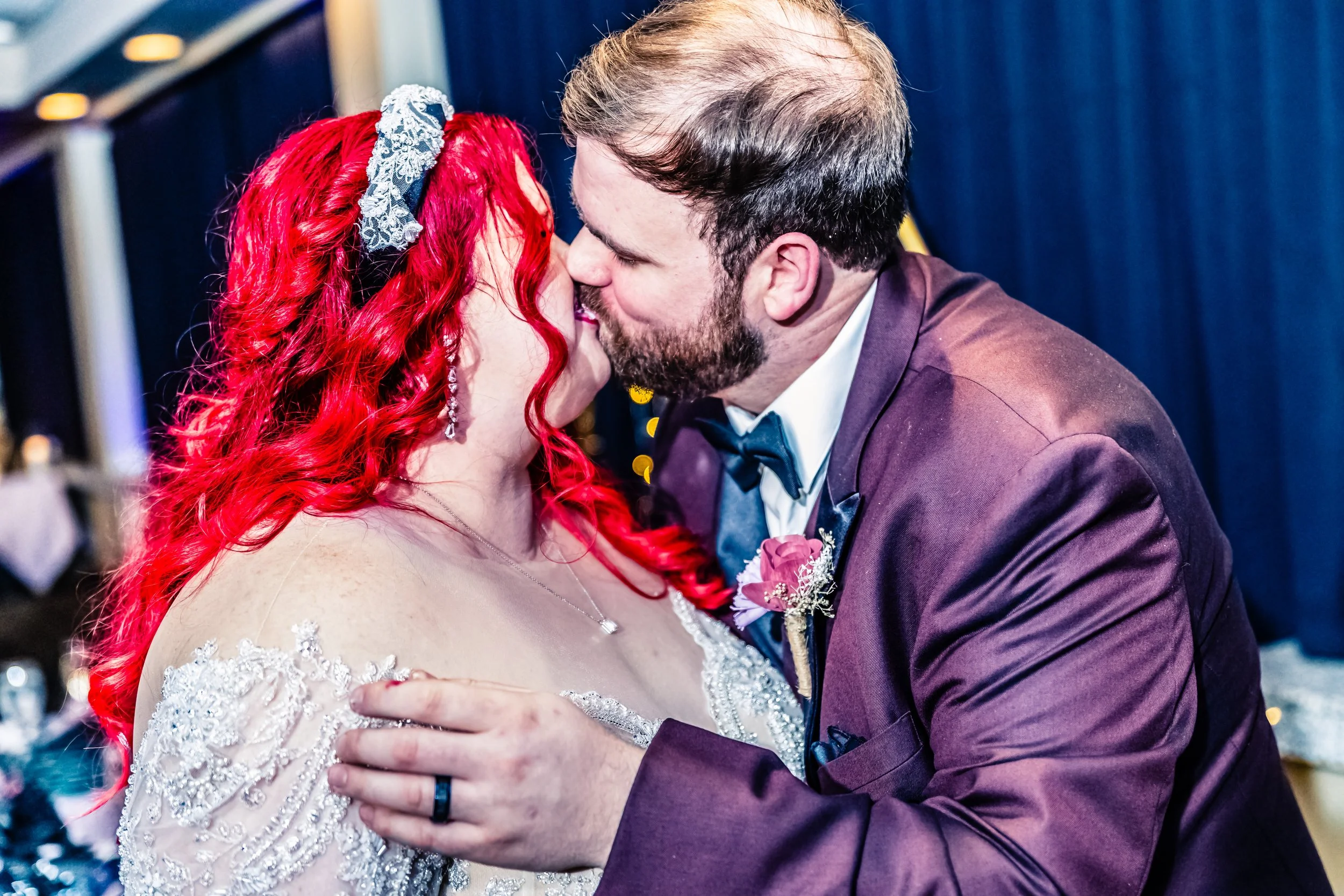 A couple in wedding attire sharing a kiss at their wedding reception, with a woman having bright red hair and a man with a beard, wearing a tuxedo with a purple jacket and a boutonniere.