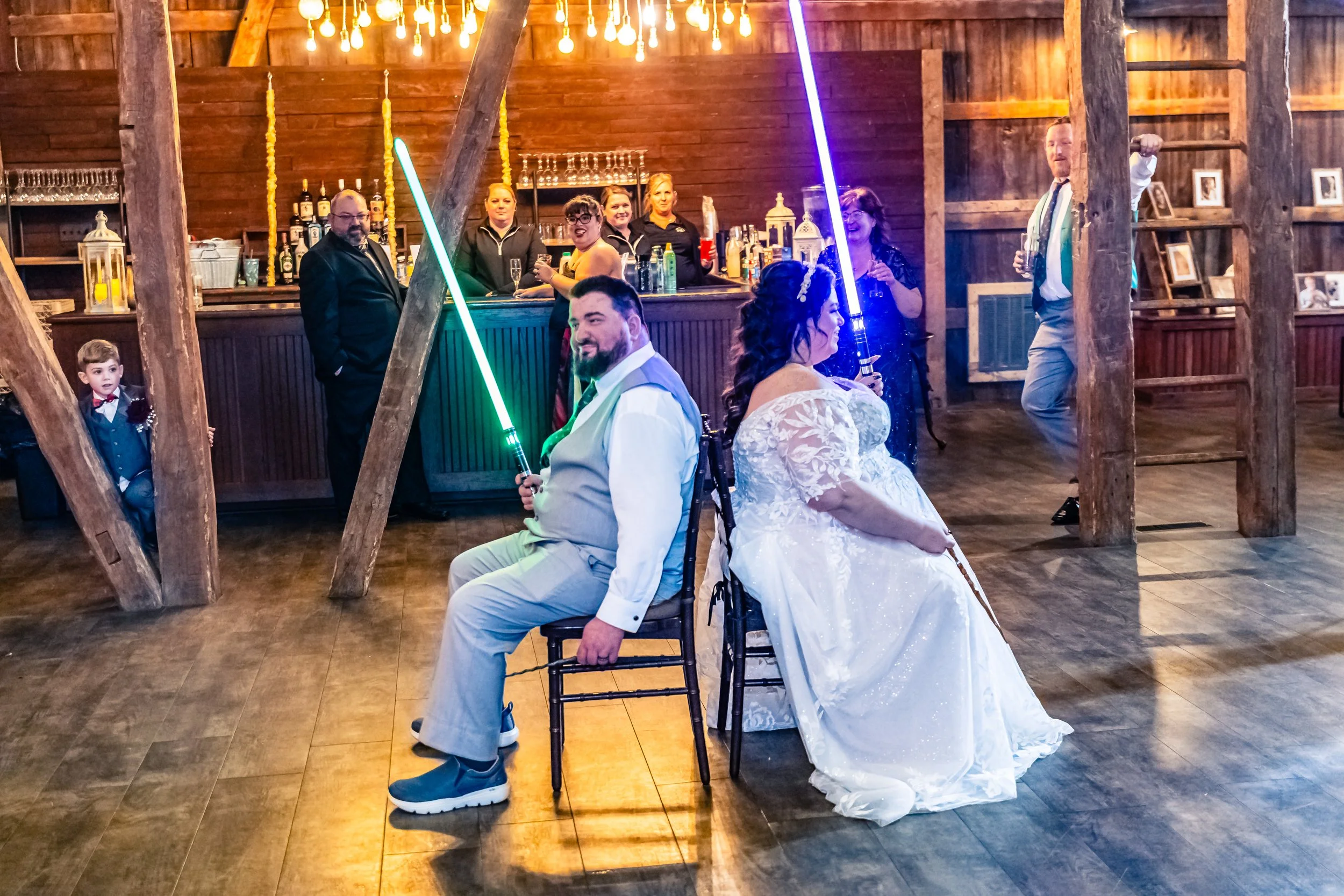 A wedding reception with a bride and groom seated back-to-back, each holding a lightsaber, in a rustic wooden venue. Guests are standing and talking near a bar in the background, with warm lighting and wooden decor.