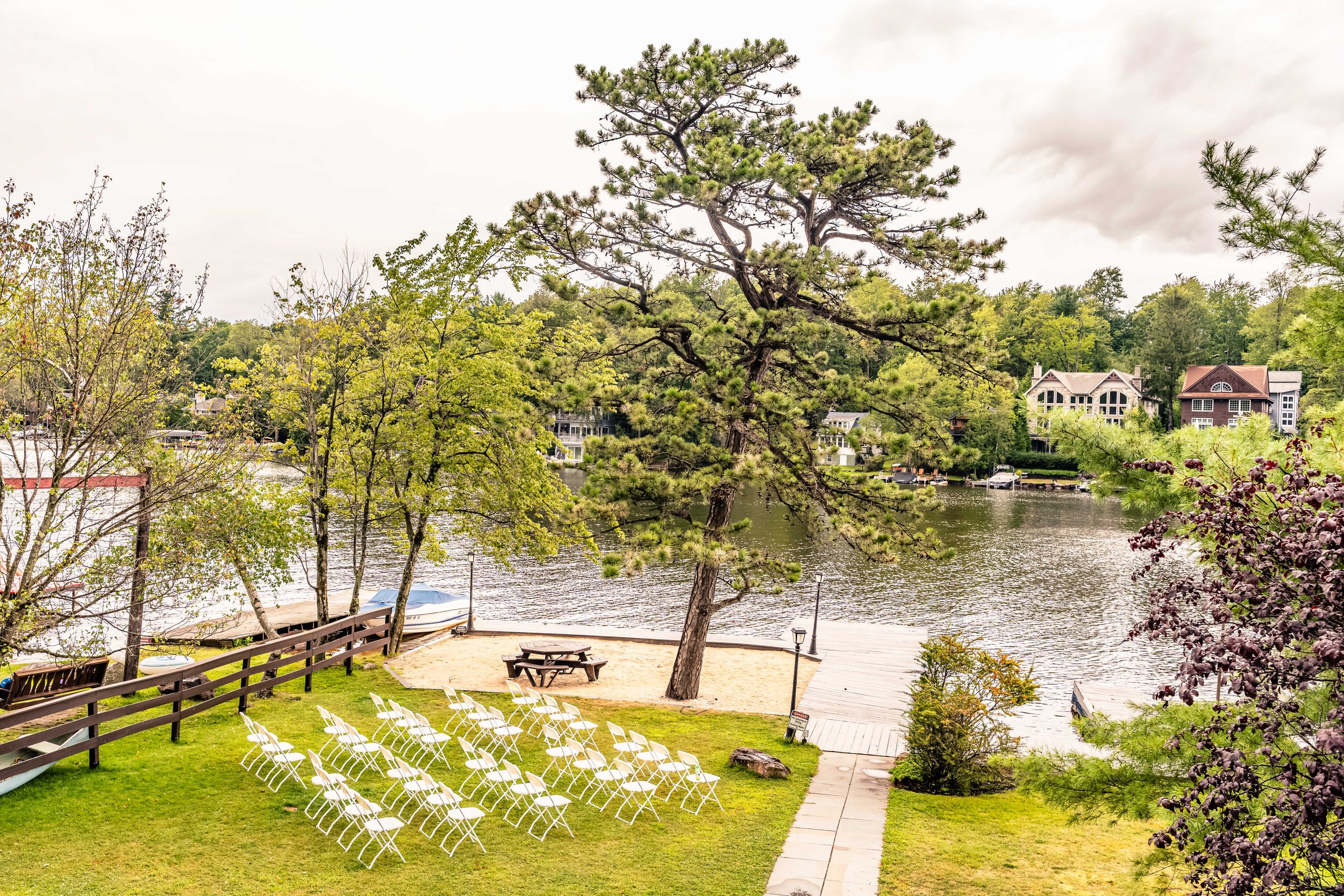 A lakeside outdoor scene with chairs arranged on a grassy lawn, a picnic table, a sandy area with a tree, a dock leading to the water, and houses in the background among trees under a cloudy sky.