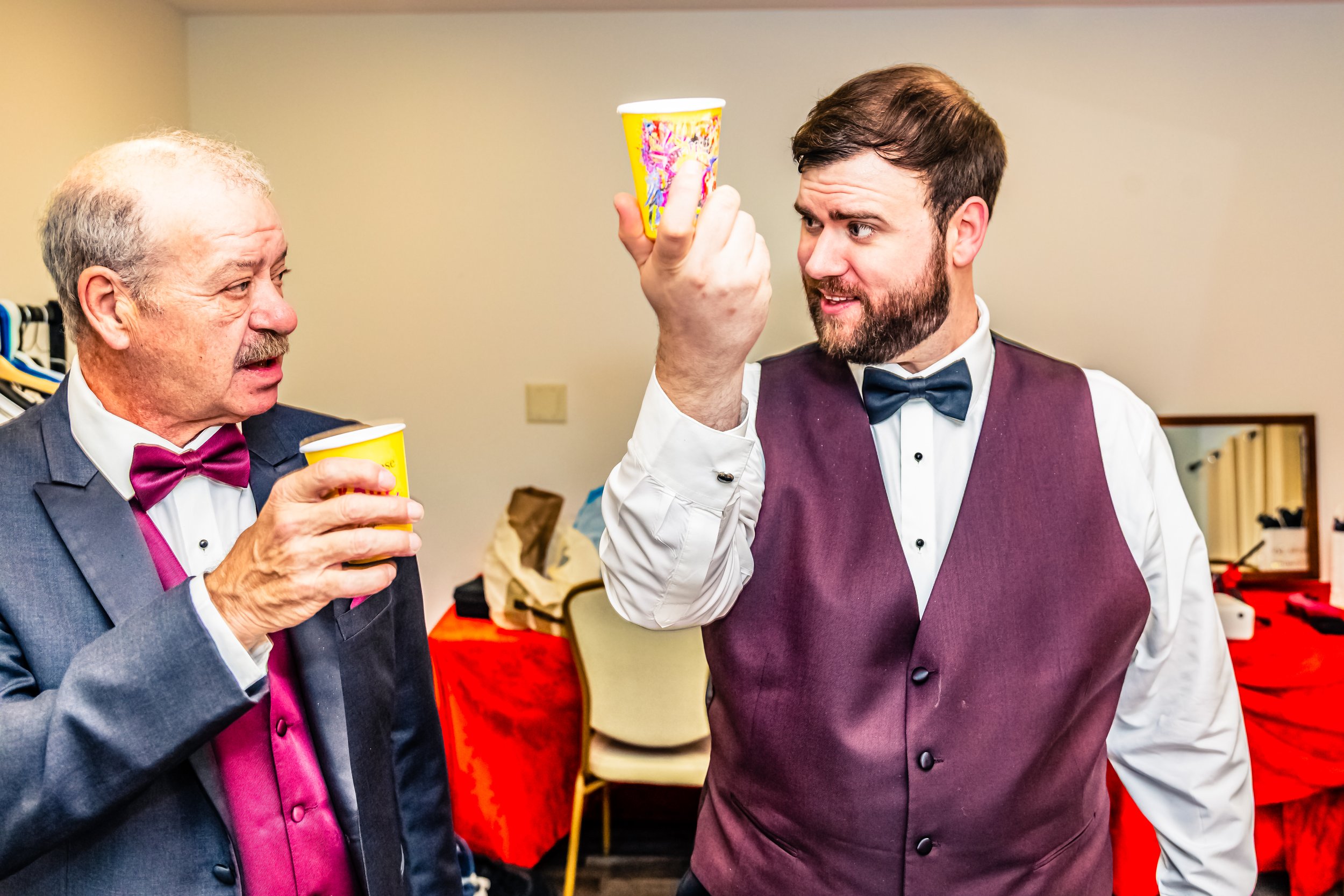Two men dressed in formal attire, holding paper cups, having a conversation in a room with a cluttered background.
