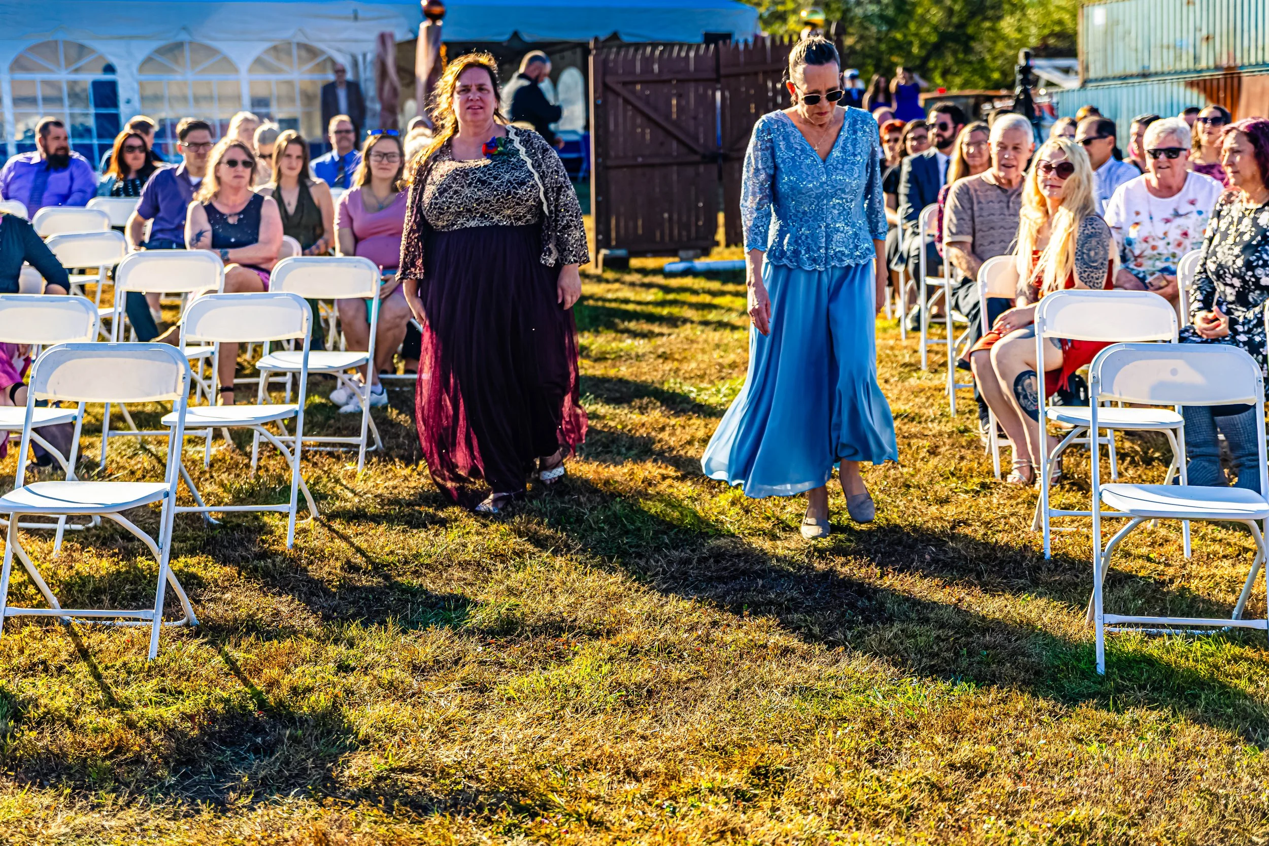 Two women walk through a sunny outdoor wedding ceremony with seated guests in the background. One woman wears a dark purple dress with gold accents, the other a light blue dress with lace top, both heads slightly bowed.
