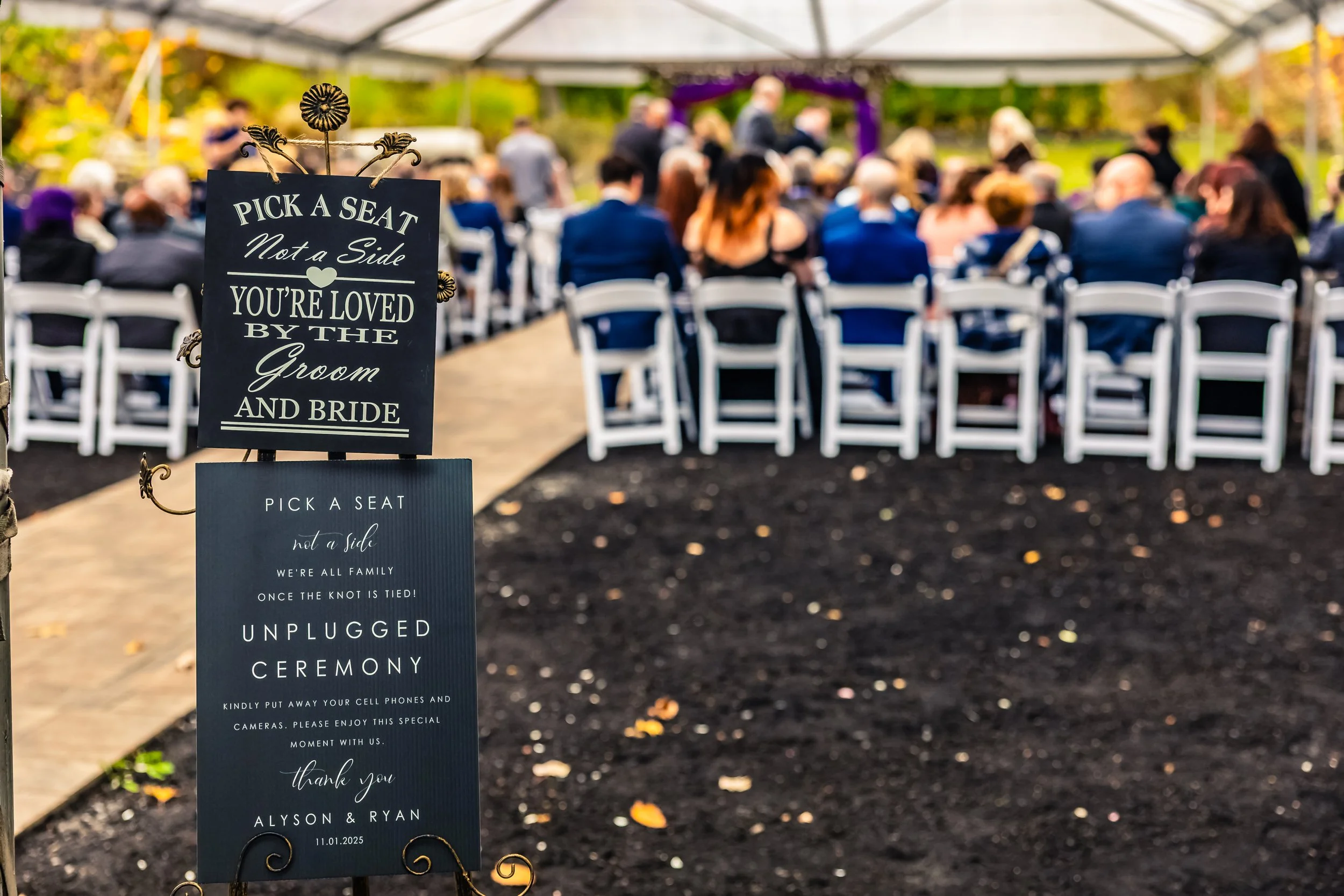 Wedding ceremony set up outdoors with white chairs arranged under a canopy, and a sign in the foreground that reads, "Pick a seat, not a side, you're loved by the groom and bride," with instructions for guests to pick a seat and unplug devices during