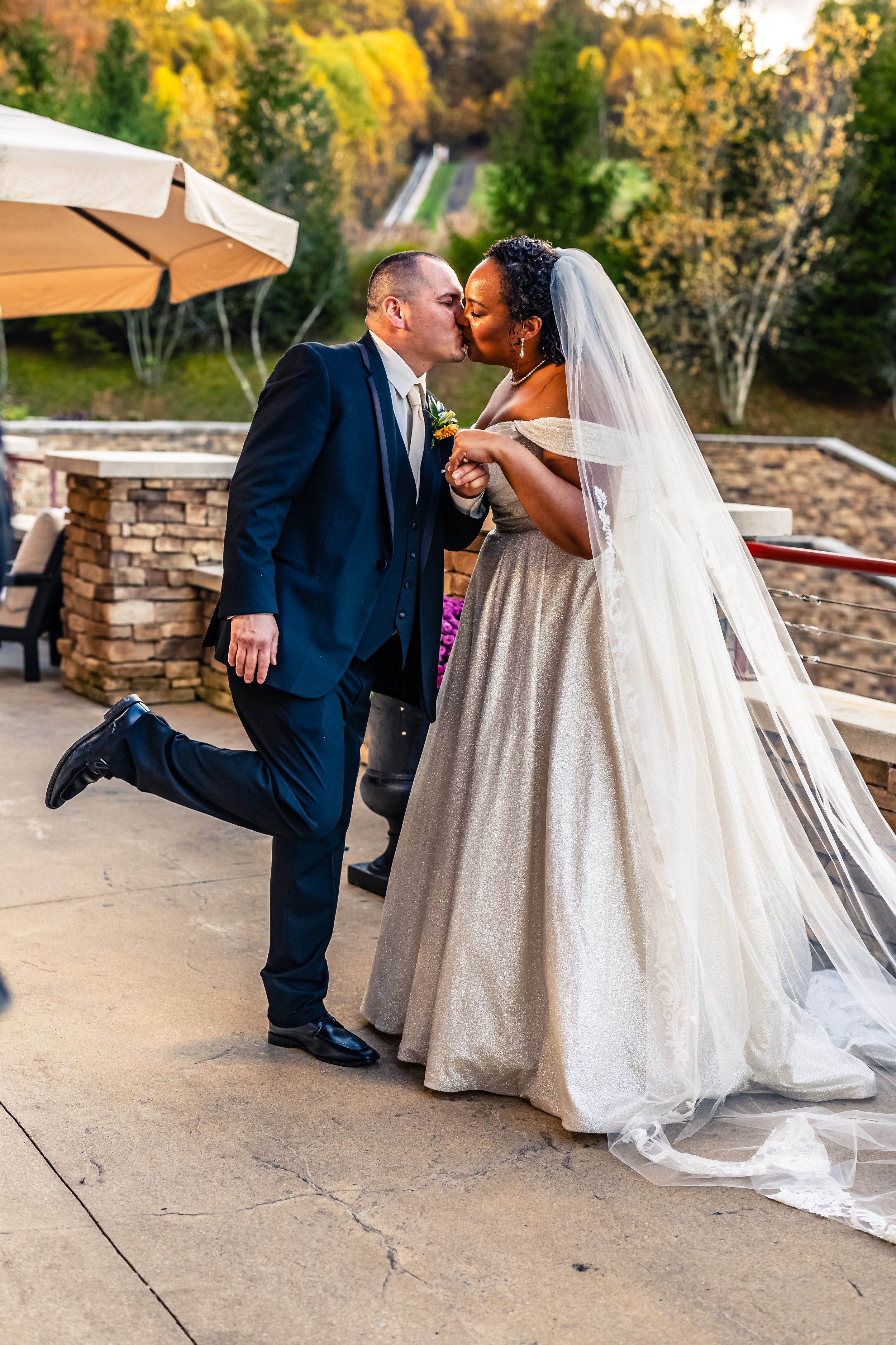 A newlywed couple sharing a kiss outdoors, with the groom kneeling on one knee while holding the bride's hand, during sunset in a scenic setting with trees and hills in the background.