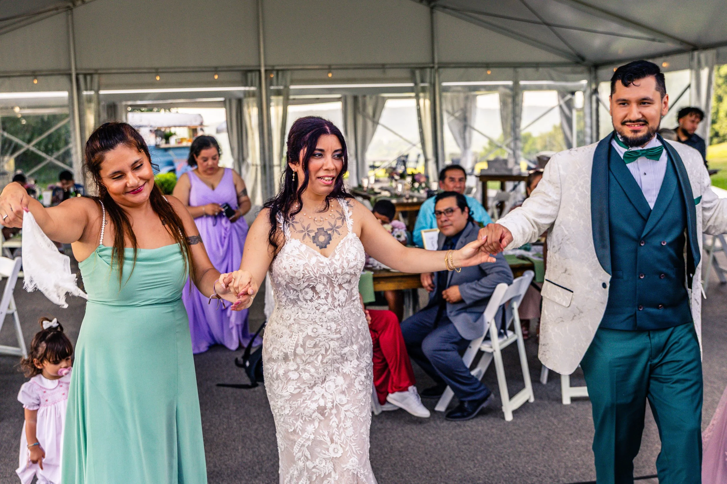 People dancing at a wedding reception inside a large white event tent, with some guests sitting at tables in the background.