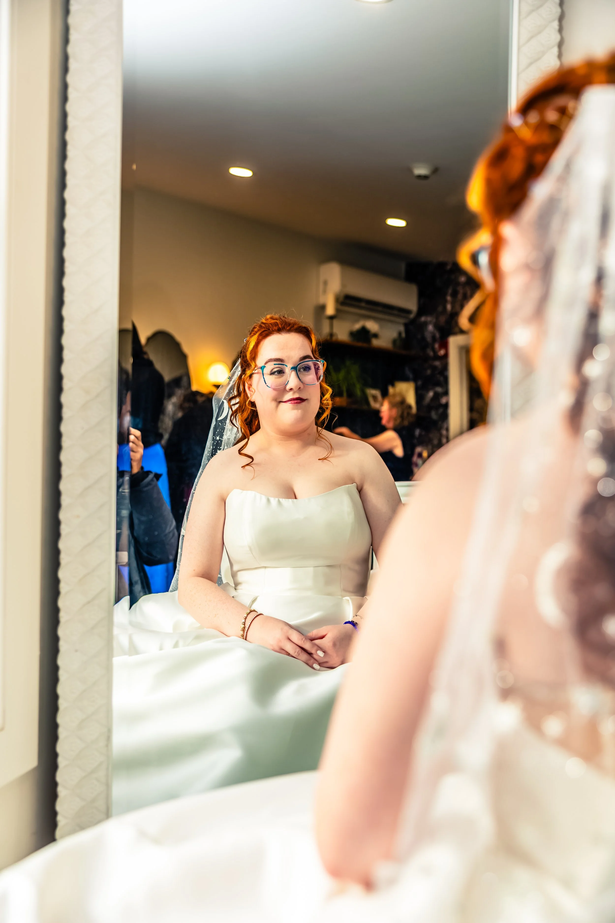 A bride with red hair and glasses looking at herself in a mirror before her wedding, wearing a strapless white wedding gown.