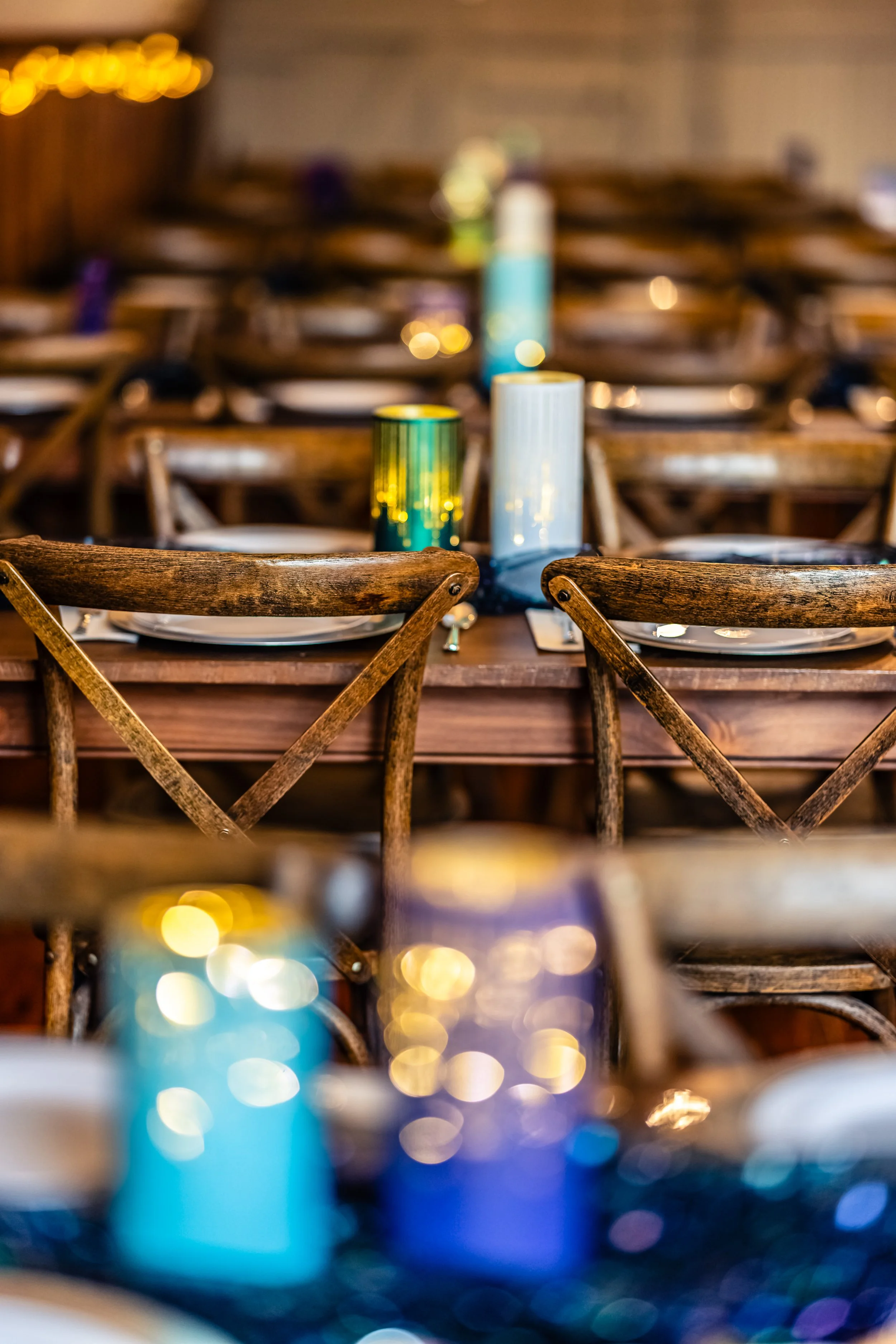 A dining table set for an event with candles and plates, surrounded by wooden chairs, in a warmly lit room.