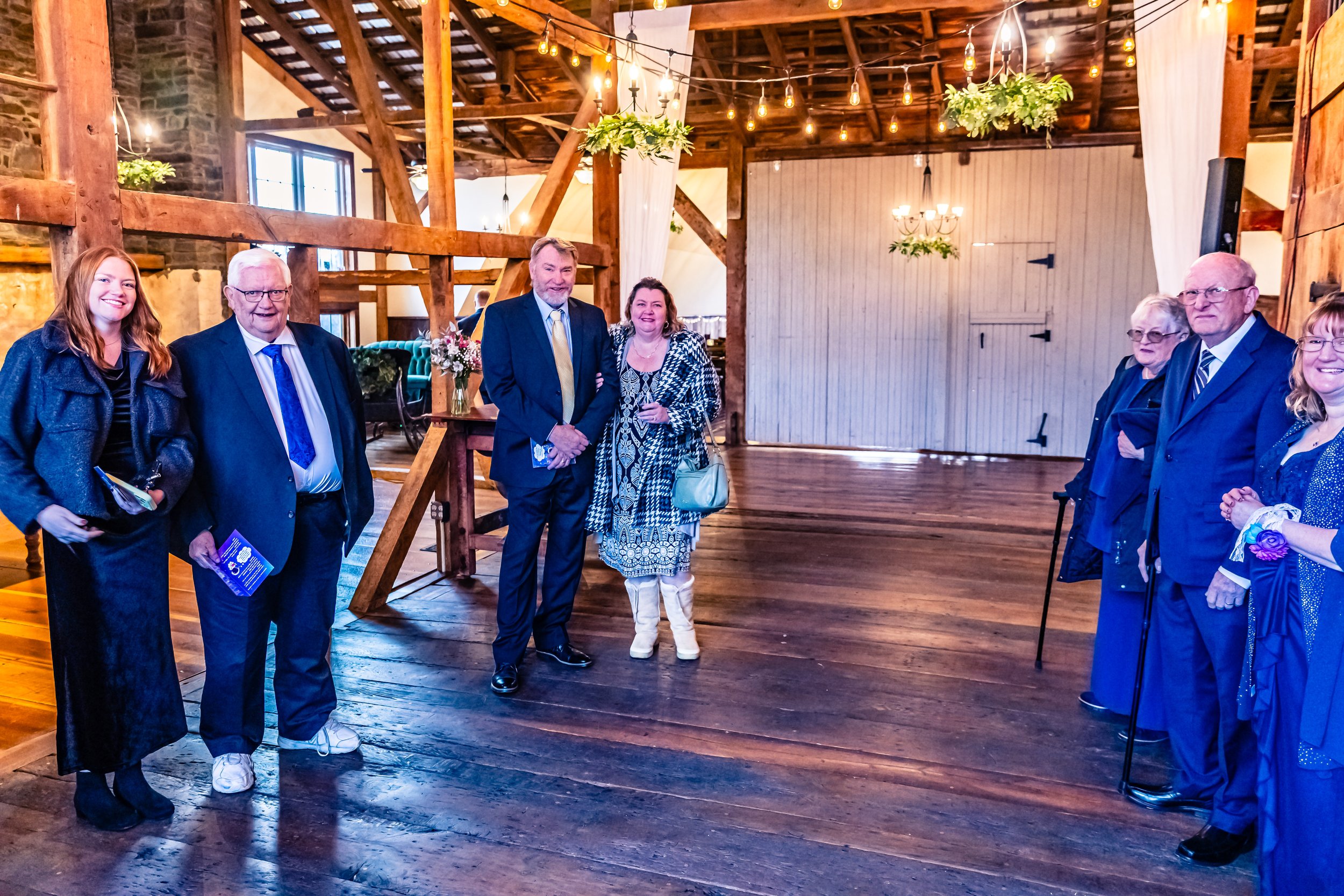 A group of people dressed in formal attire standing inside a rustic wooden venue with wood beams, chandeliers, and string lights.
