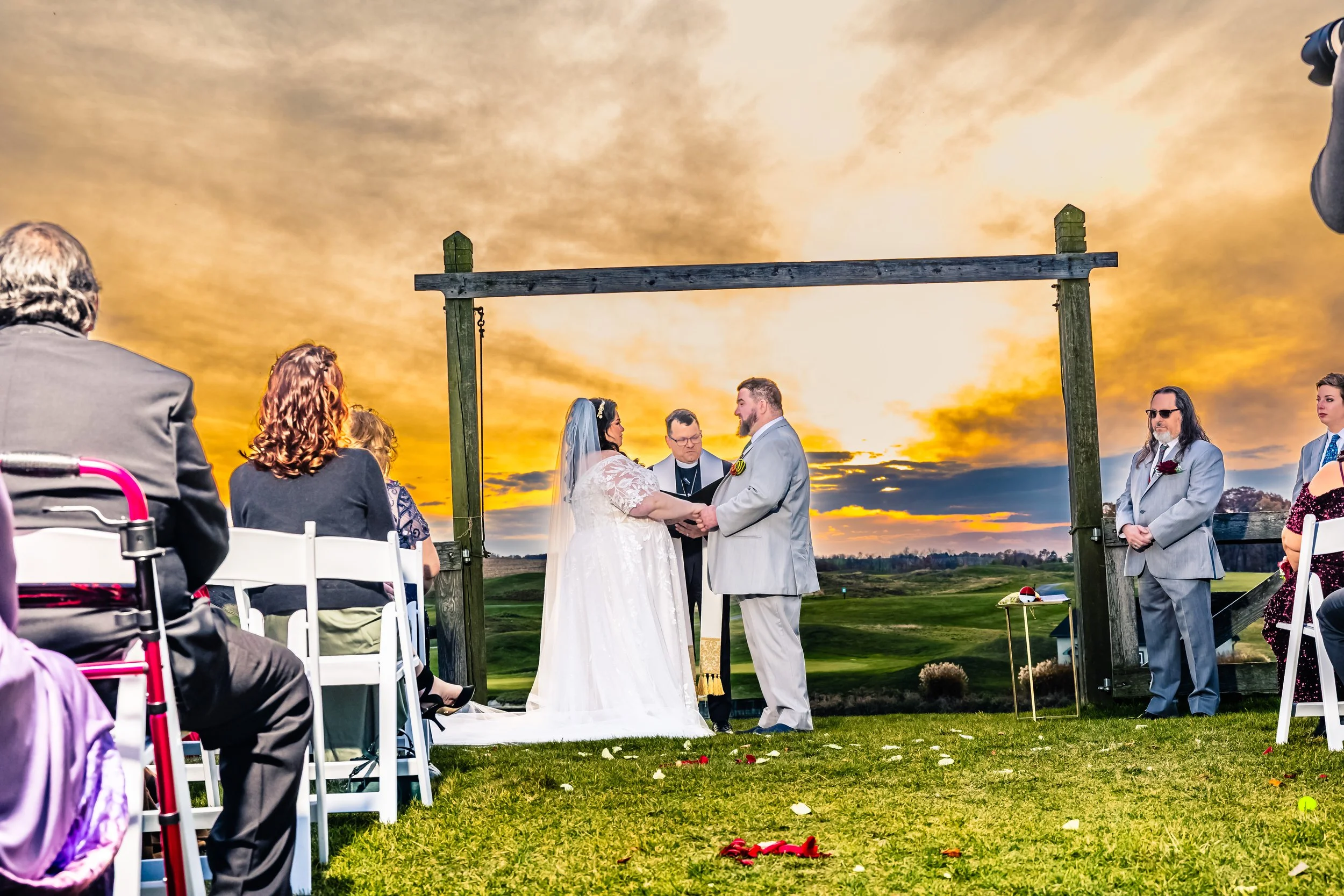 A wedding ceremony taking place outdoors at sunset, with the bride and groom holding hands and standing under a wooden arch, surrounded by guests seated on white chairs, on a grassy field with a scenic view of the sky and clouds.