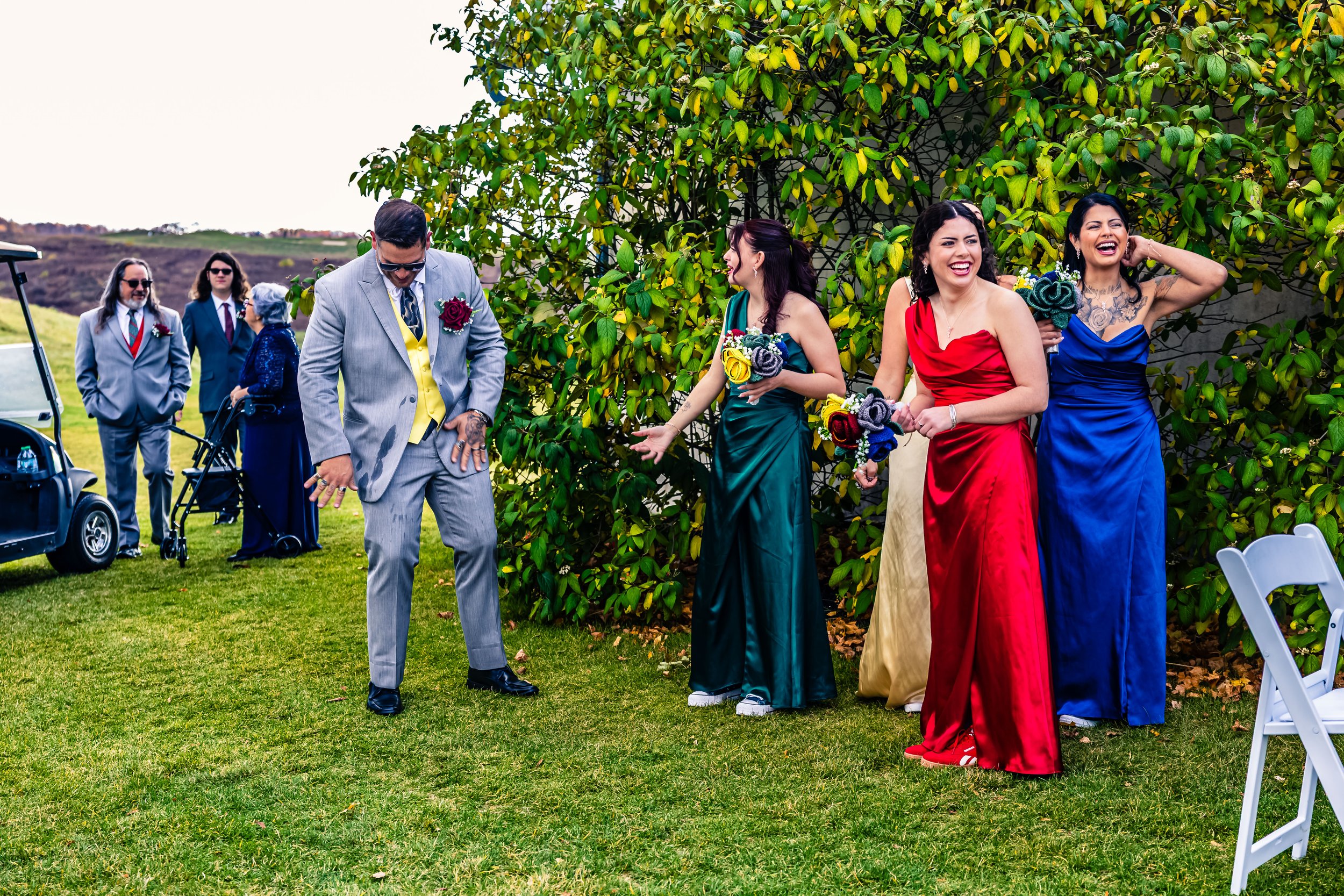 A group of wedding guests, including groomsmen and bridesmaids, standing on a lawn near a bush. The bridesmaids are wearing colorful dresses and holding bouquets, while the groomsmen are in suits. The scene is cheerful and relaxed.