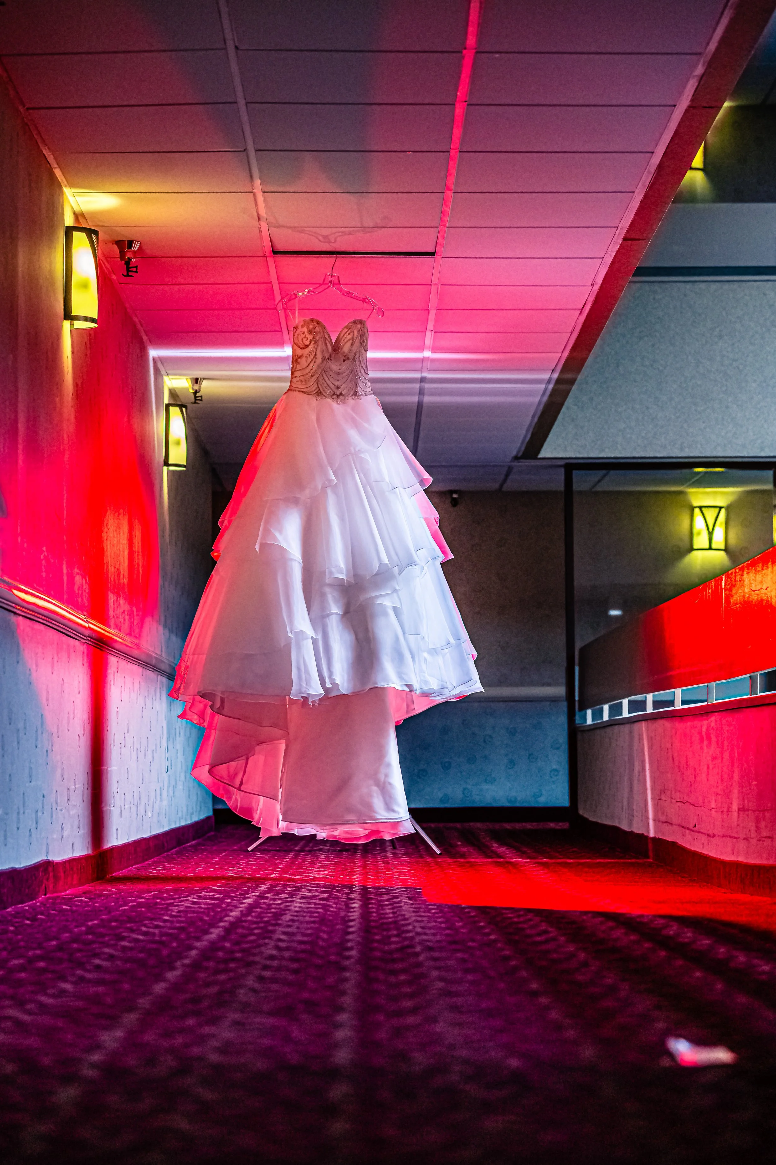 A white wedding dress hanging on a hanger from the ceiling in a corridor with red and yellow lights.