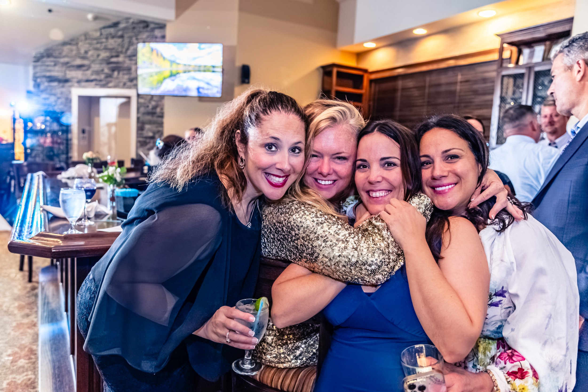 Group of four women smiling and hugging at a social event, with a decorated restaurant or banquet hall background.