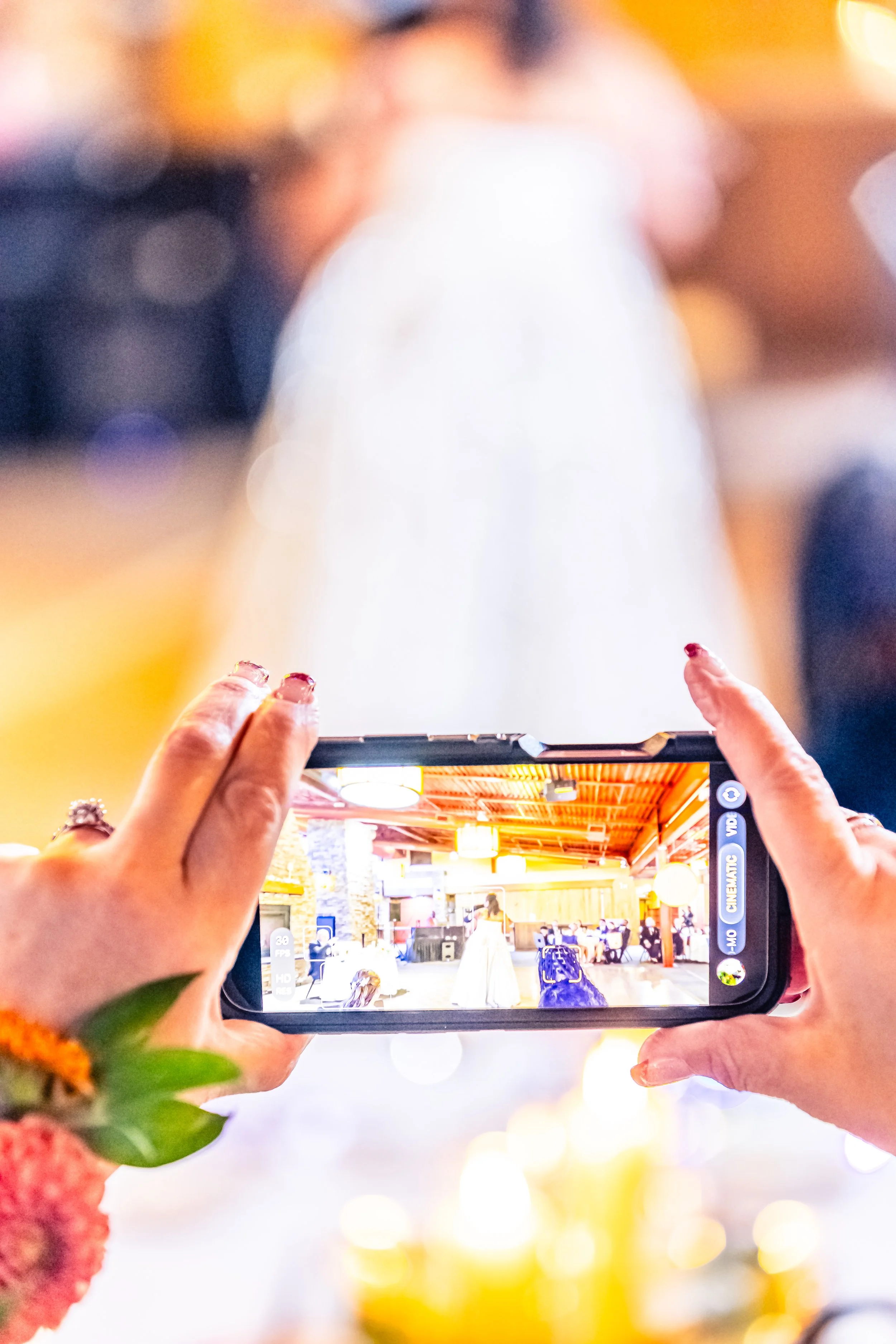 Person taking a photo of a bride in a white dress inside a venue with wooden ceilings and warm lighting.
