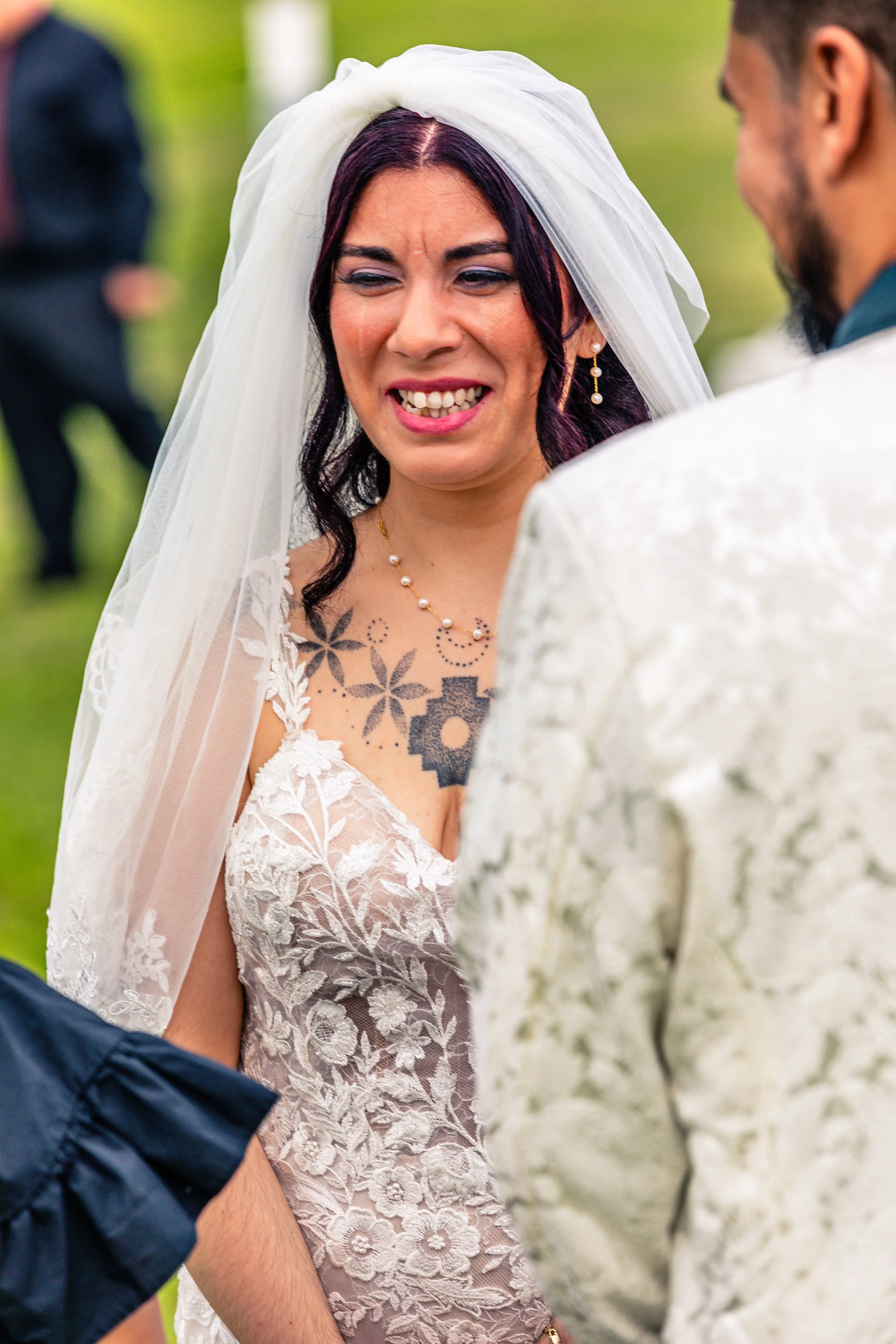 A woman with purple hair and tattoos getting married outside, wearing a white veil and a lace wedding dress, smiling while exchanging vows.