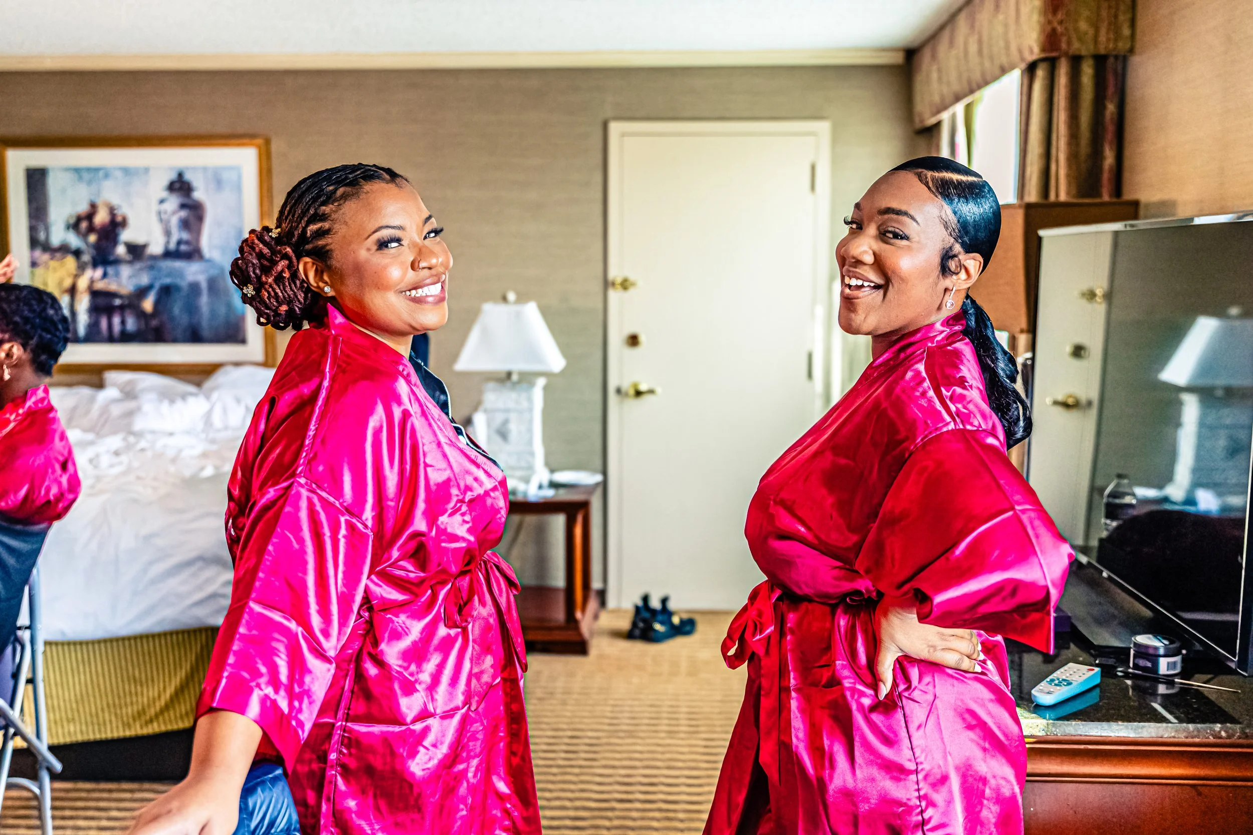 Two smiling women in pink satin robes standing in a bedroom, looking at the camera.