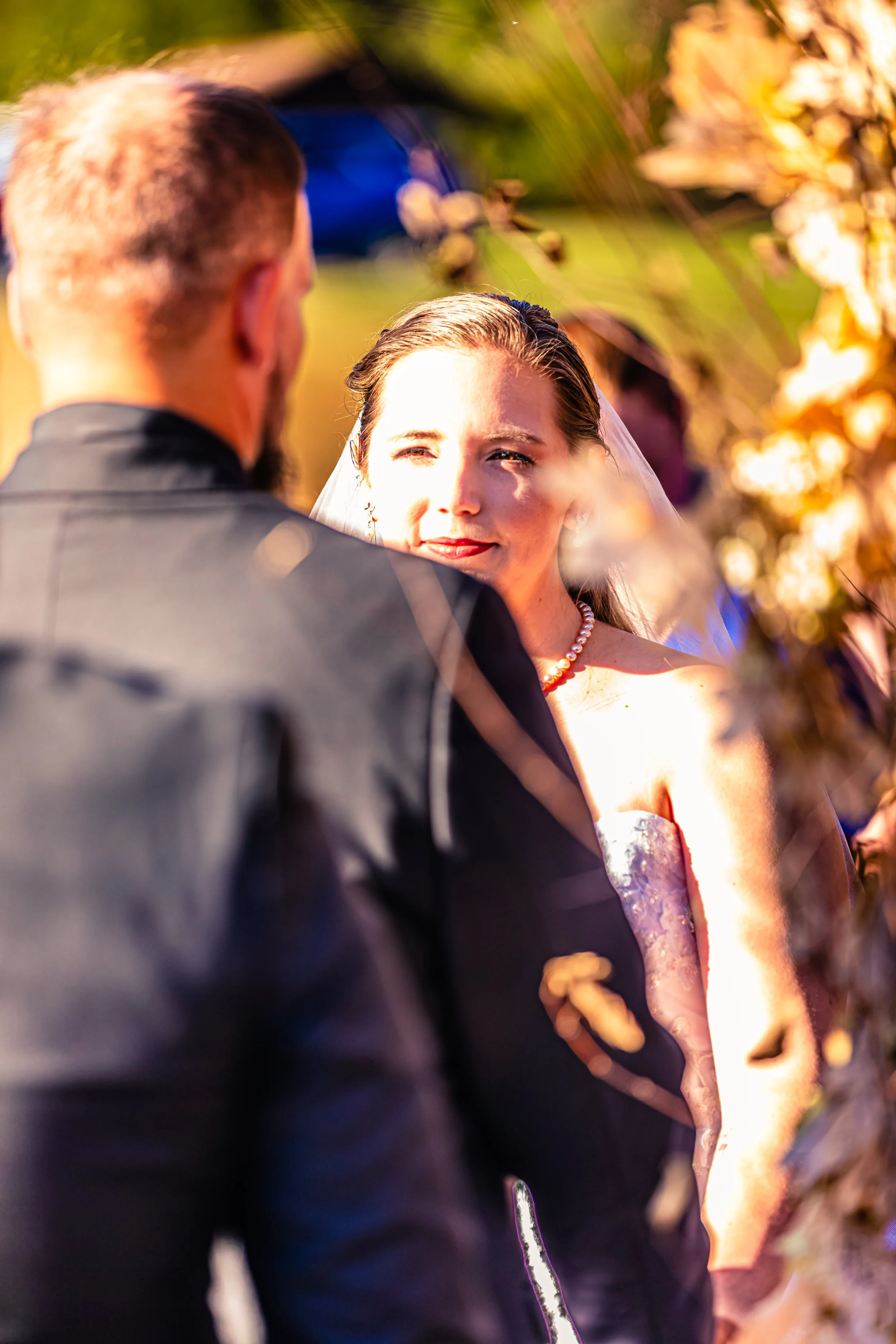 A bride and groom standing outdoors, with the bride looking at the groom as he faces her, surrounded by greenery and warm sunlight.