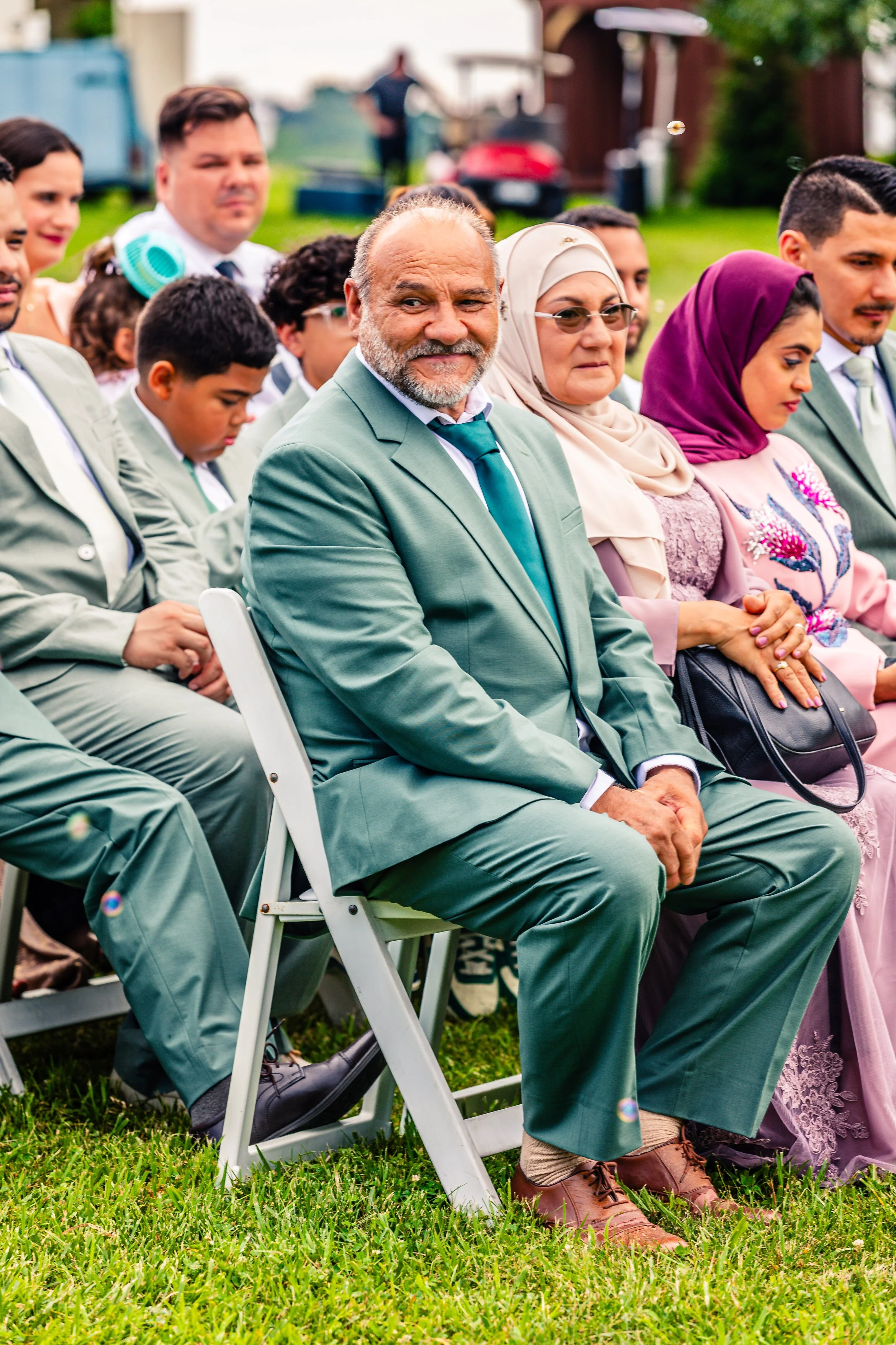 Group of people dressed formally, seated outdoors on grass, attending a ceremony or event.