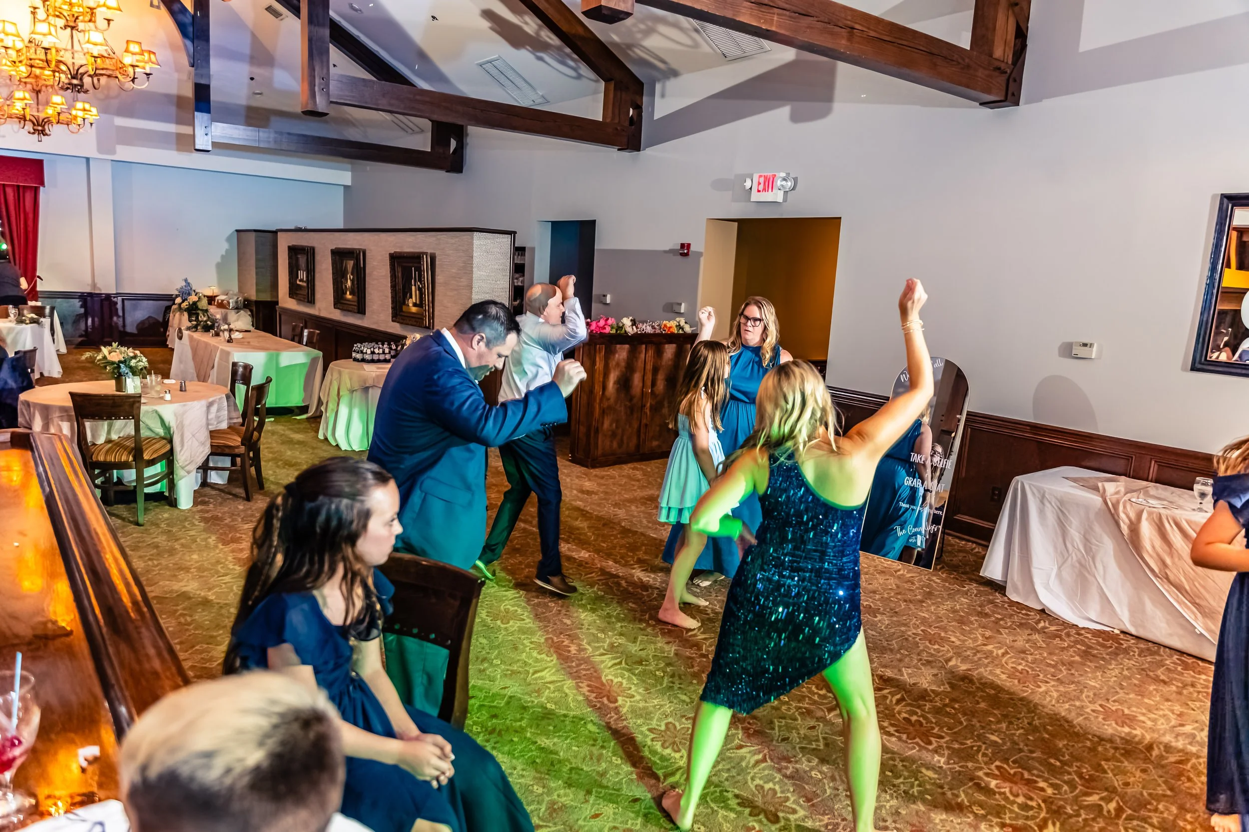 People dancing and socializing in a decorated event hall with tables, chairs, and floral centerpieces.