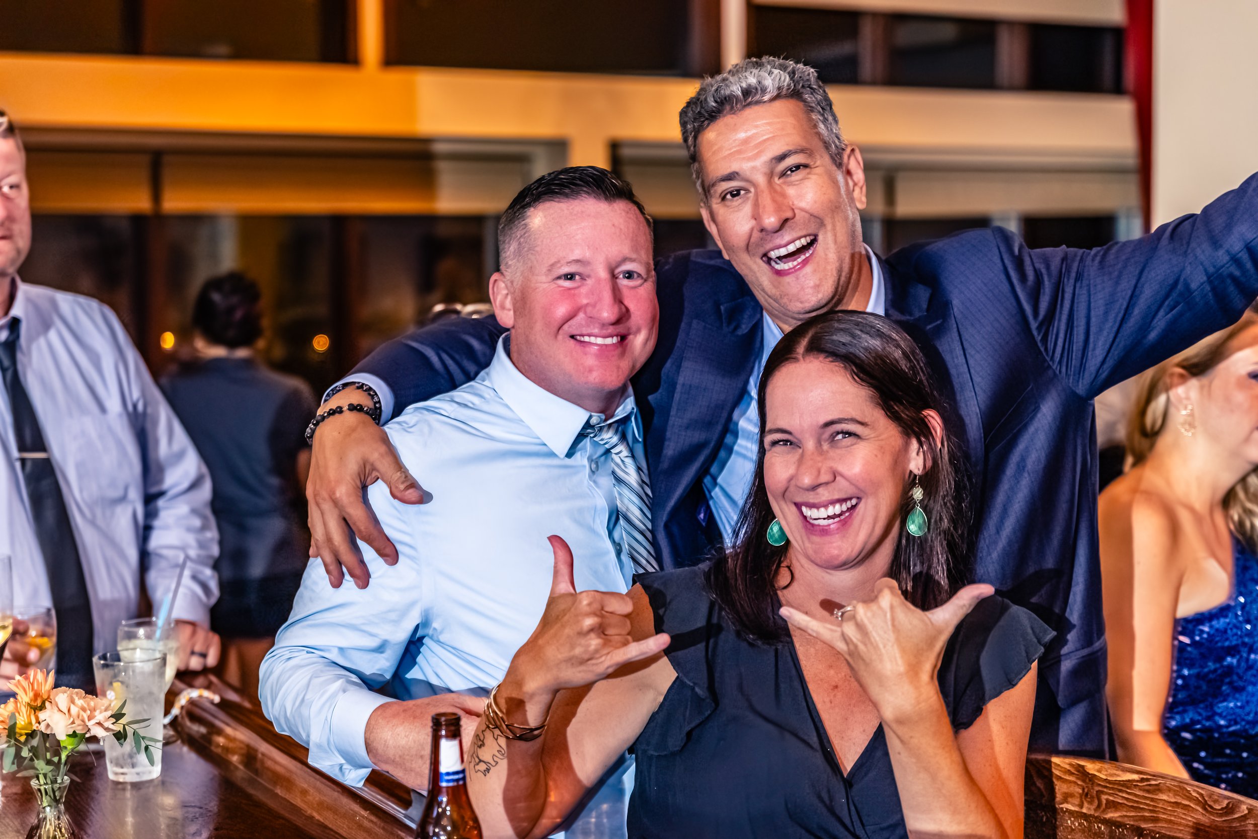 Three people smiling and posing at a lively party or celebration, with a man on the right raising his arm, and a woman in front making a rock and roll hand gesture.