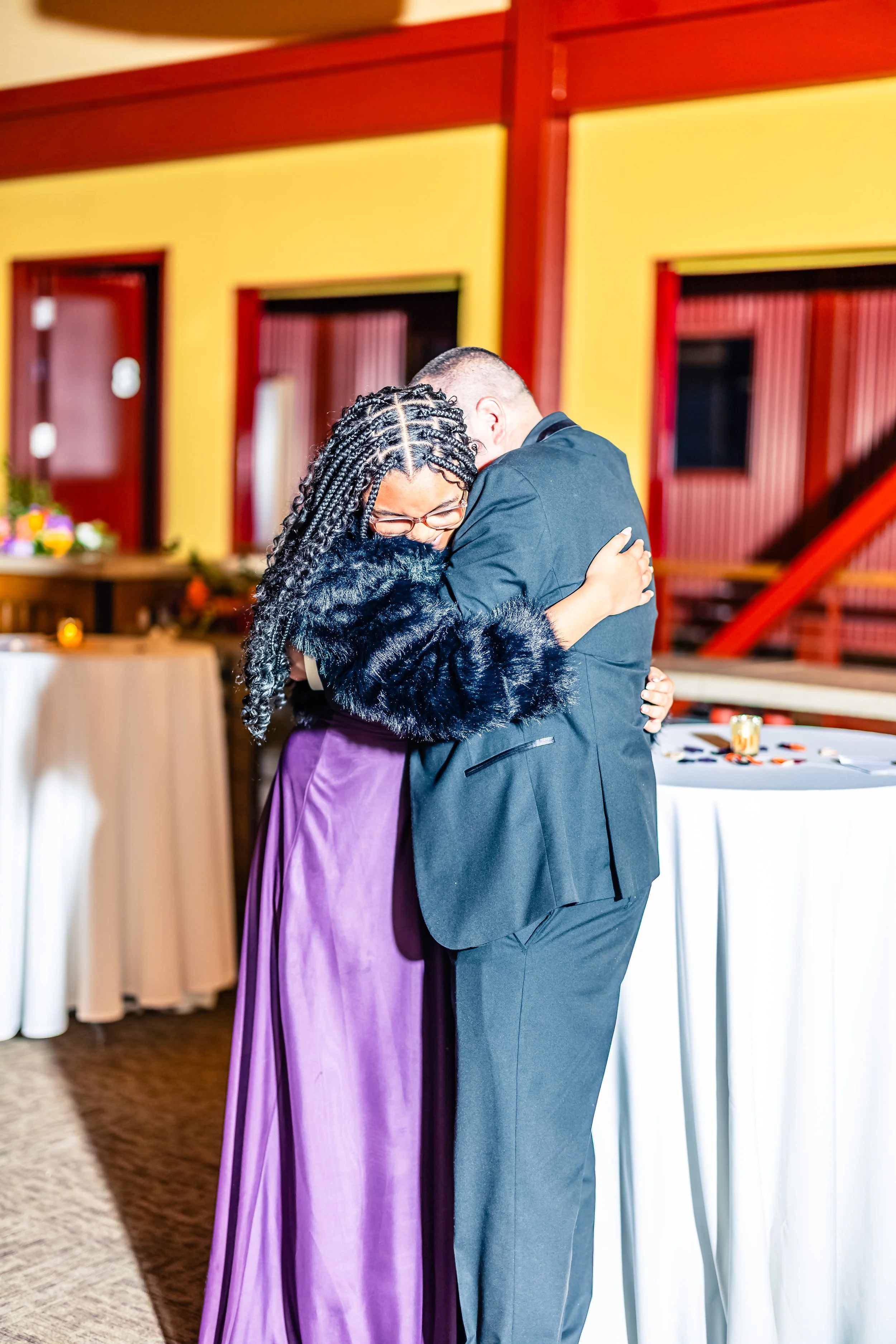 Two people hugging, one woman with braids wearing a purple dress and a faux fur black jacket, and a man in a black suit, inside a warmly lit decorated room.