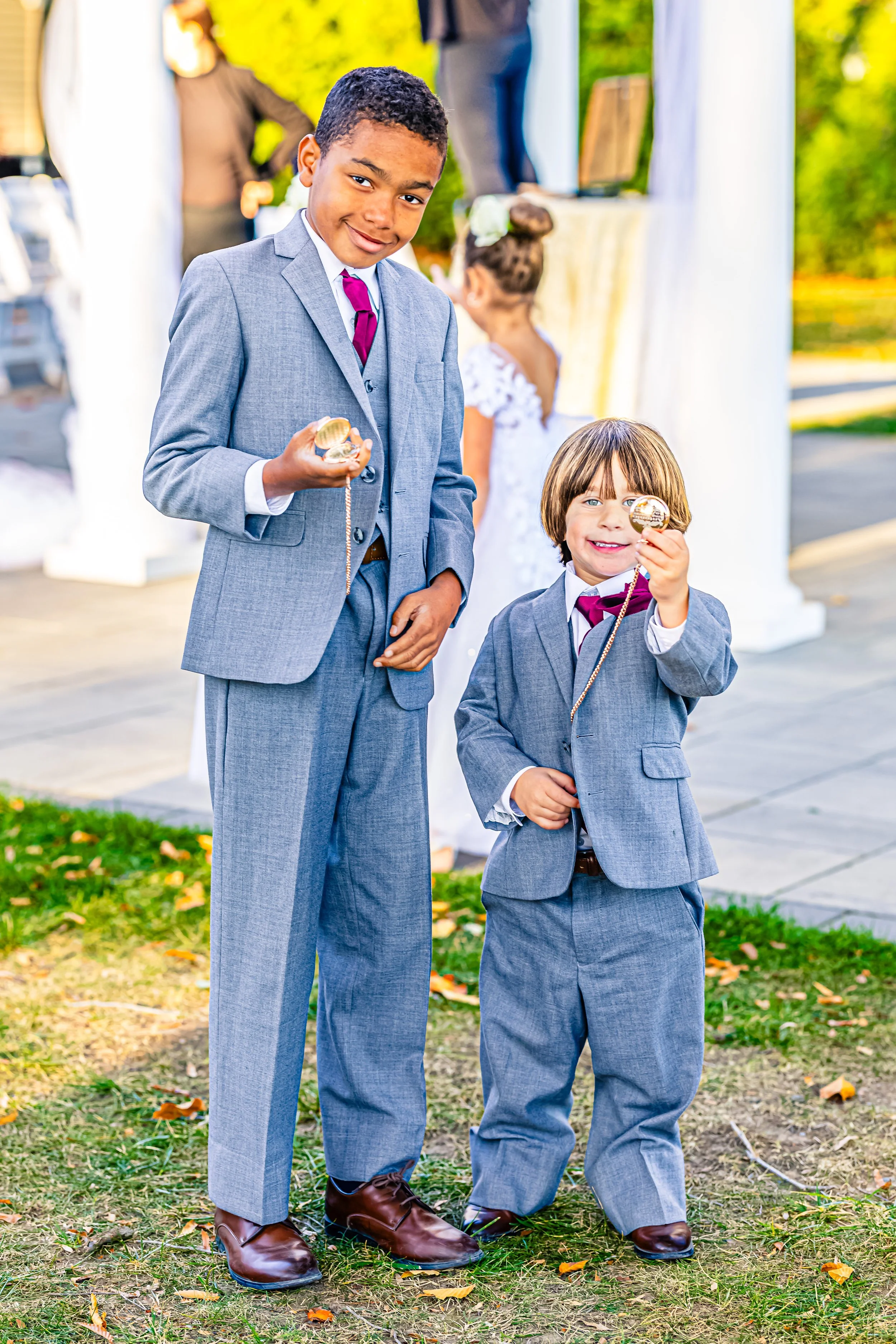 Two boys in gray suits standing outdoors, holding gold pocket watches, smiling, at a formal event with white columns and a woman in a white dress in the background.