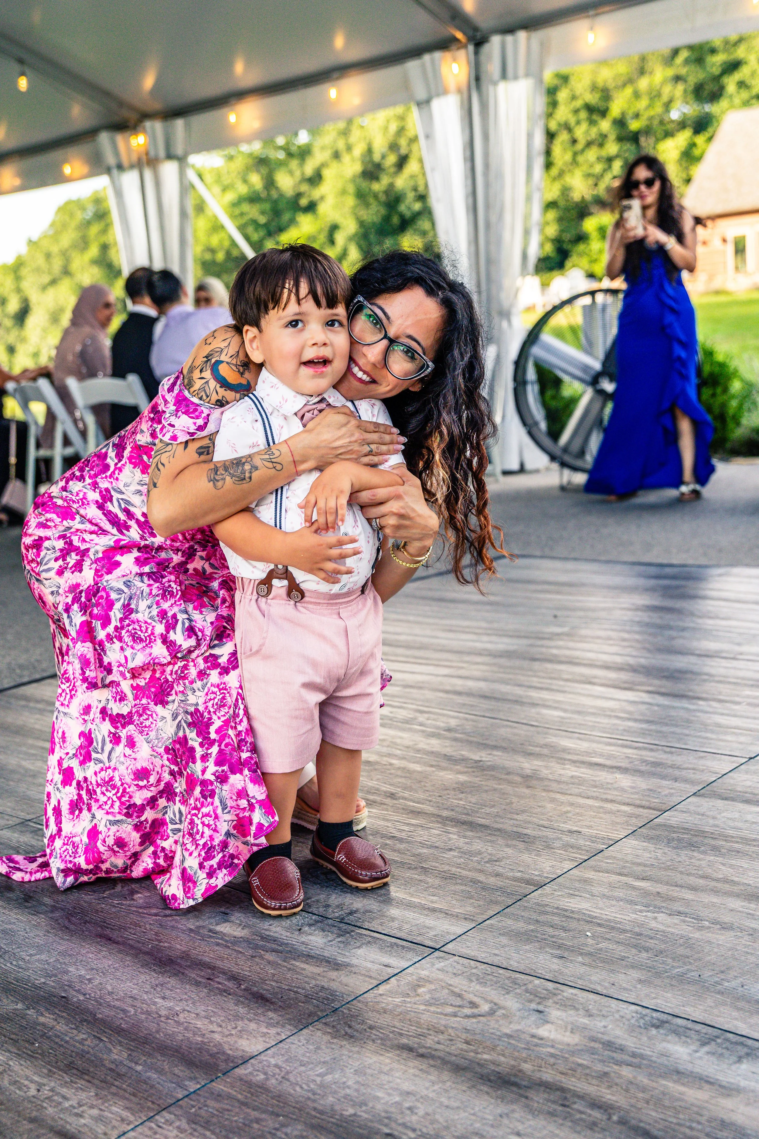 A woman with glasses and dark curly hair hugging a young boy at an outdoor event under a tent.