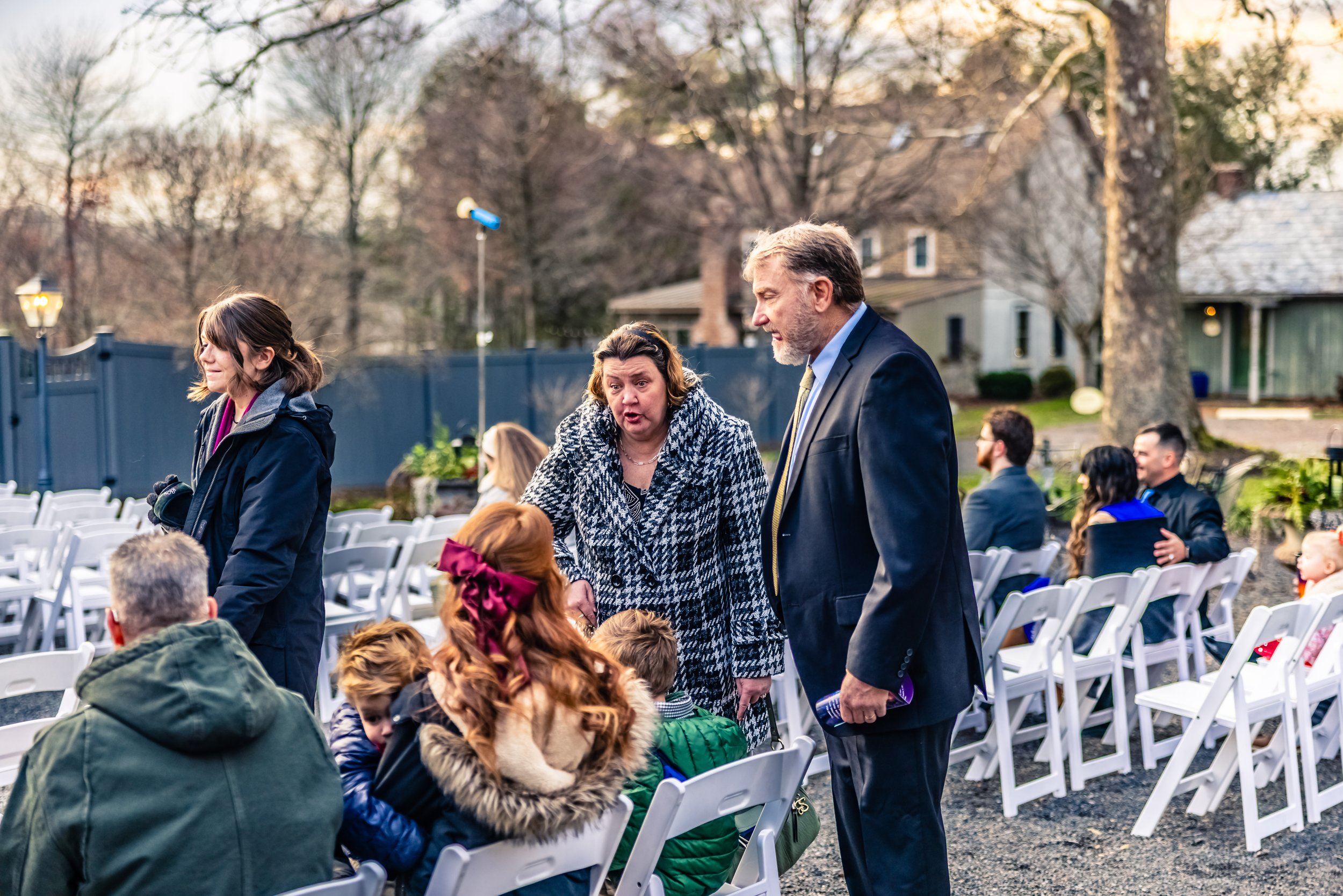 People gathered outdoors at a social event with white folding chairs, talking and interacting, with trees and residential houses in the background.