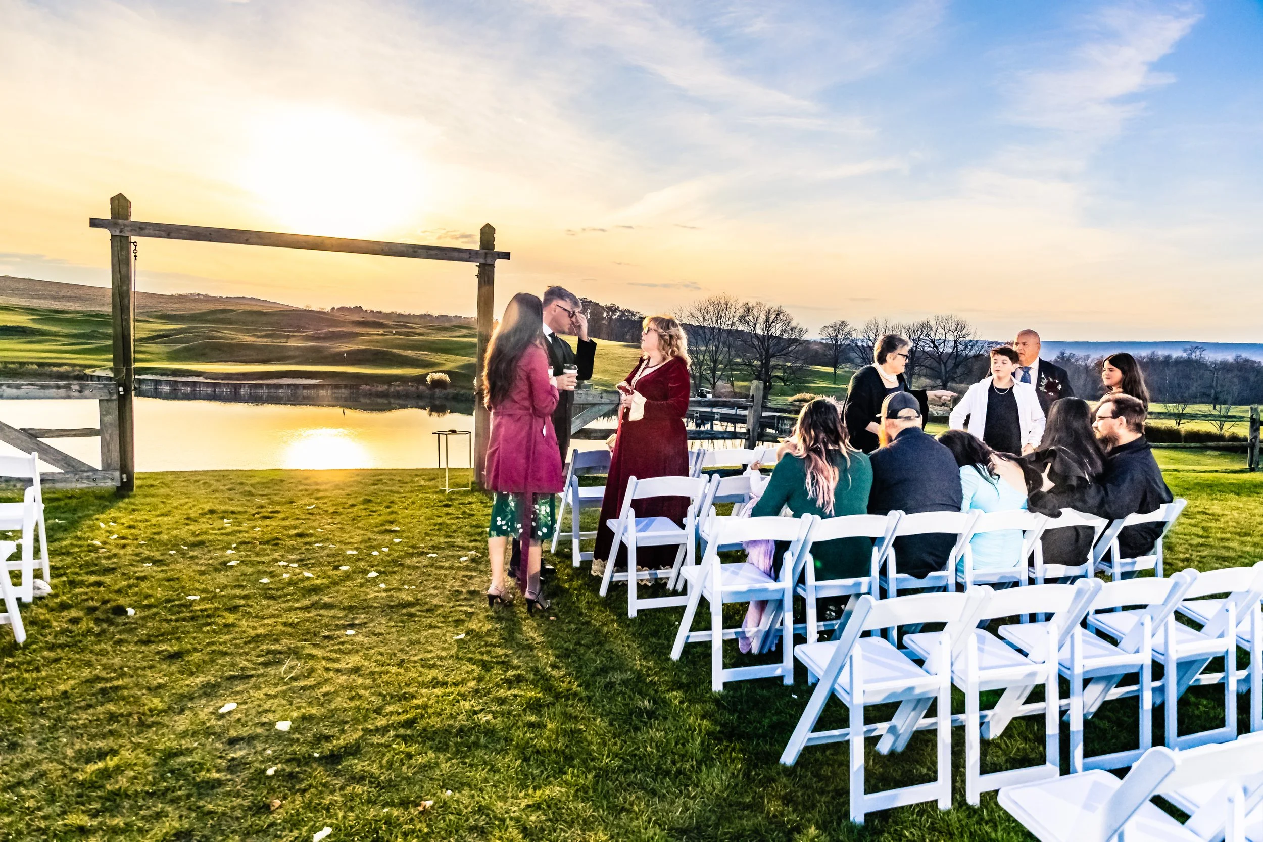 A group of people gathered outdoors near a pond at sunset, sitting on white chairs, with some standing and talking, in a scenic area with green grass, trees, and rolling hills in the background.