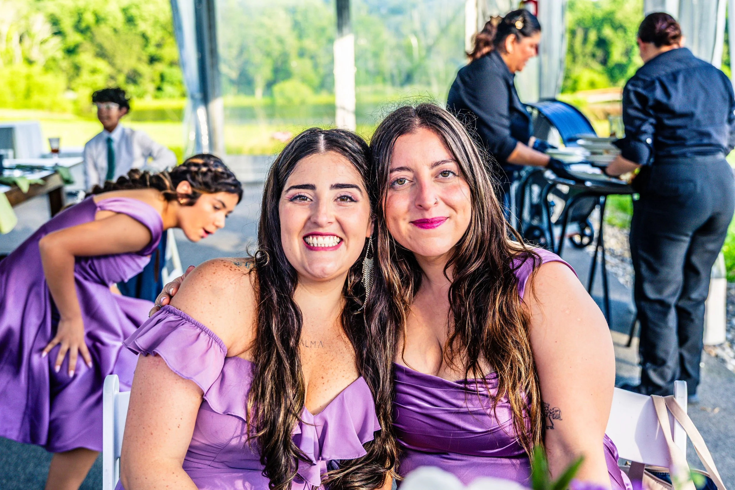 Two women smiling and wearing purple dresses, sitting at an outdoor event, with server staff and a lakeside view in the background.