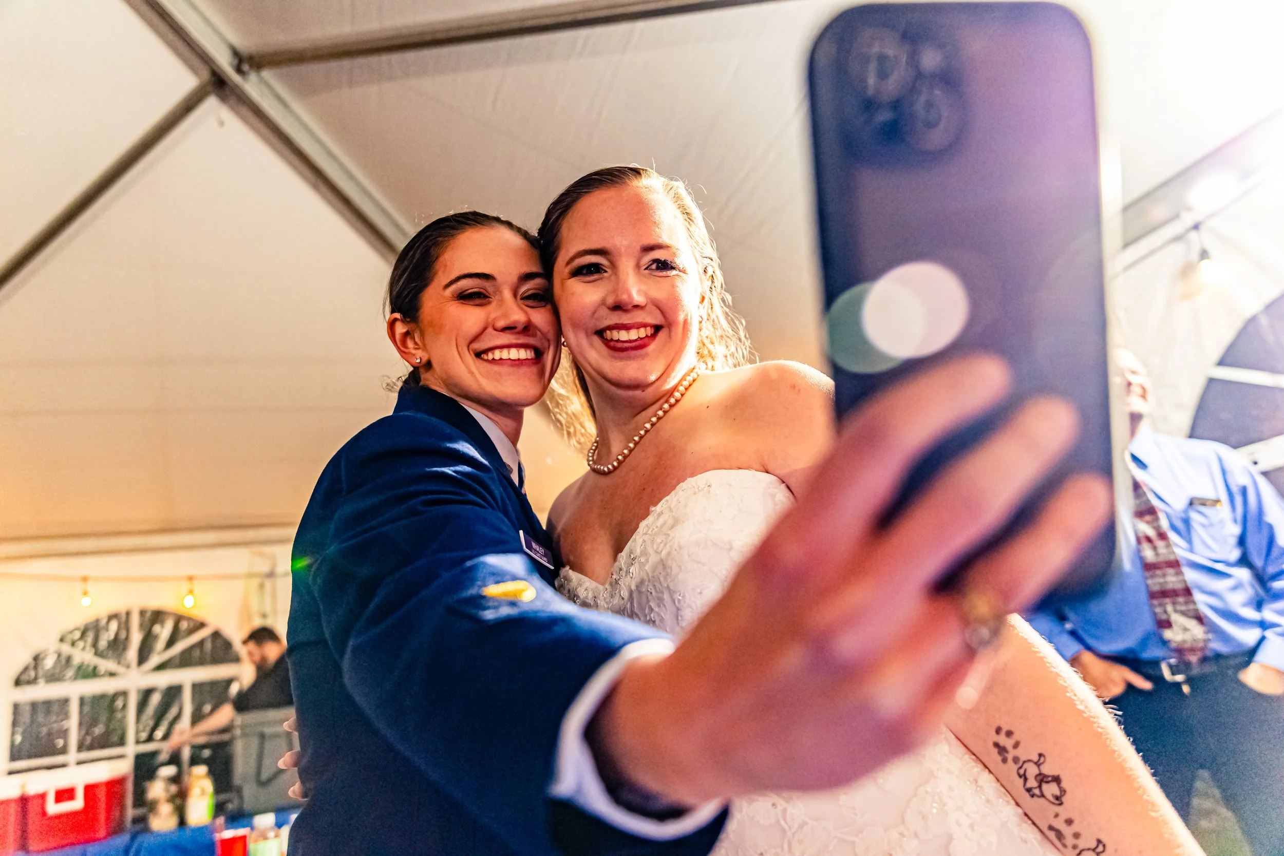 Two women in wedding attire taking a selfie inside a tent, smiling closely together, with one woman wearing a pearl necklace and the other in a navy jacket.