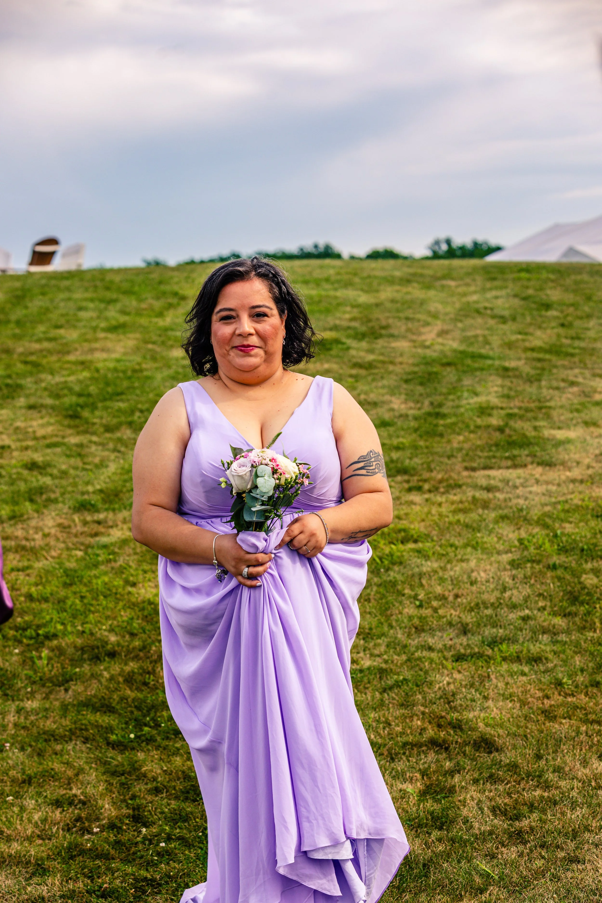 A woman in a lavender dress holding a bouquet of flowers standing on a grassy field with tents in the background.