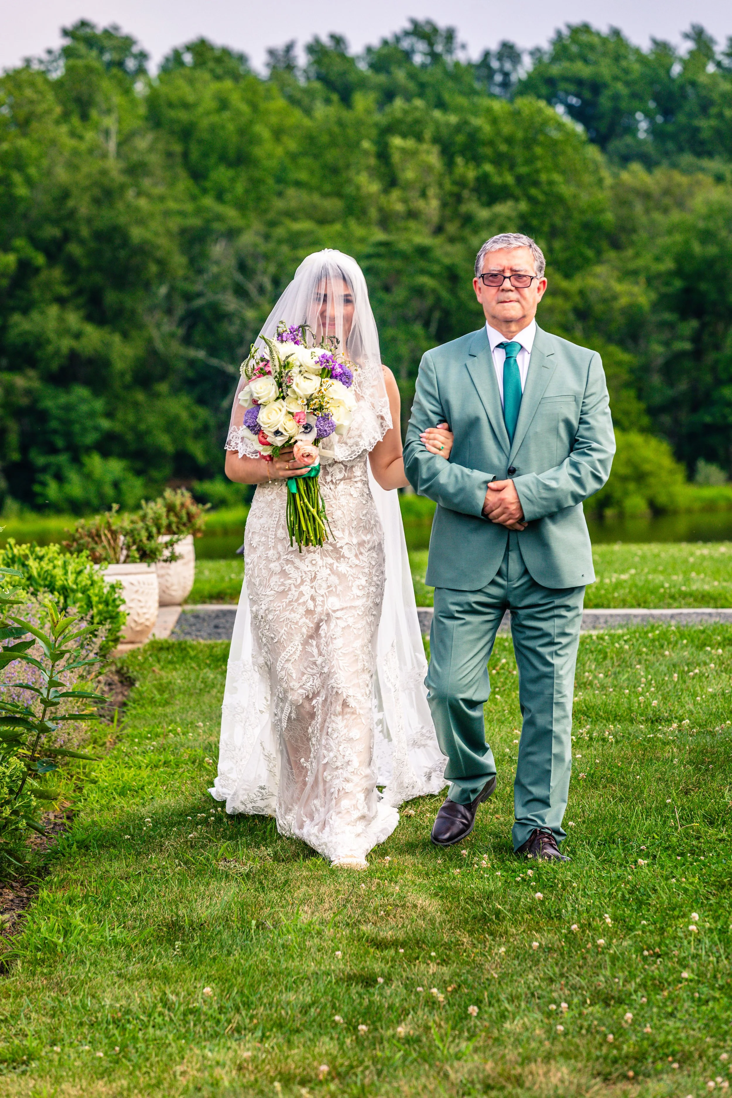 A bride in a lace wedding gown and veil holding a bouquet of white and purple flowers, walking arm-in-arm with an older man in a light green suit on a grassy lawn with trees in the background.