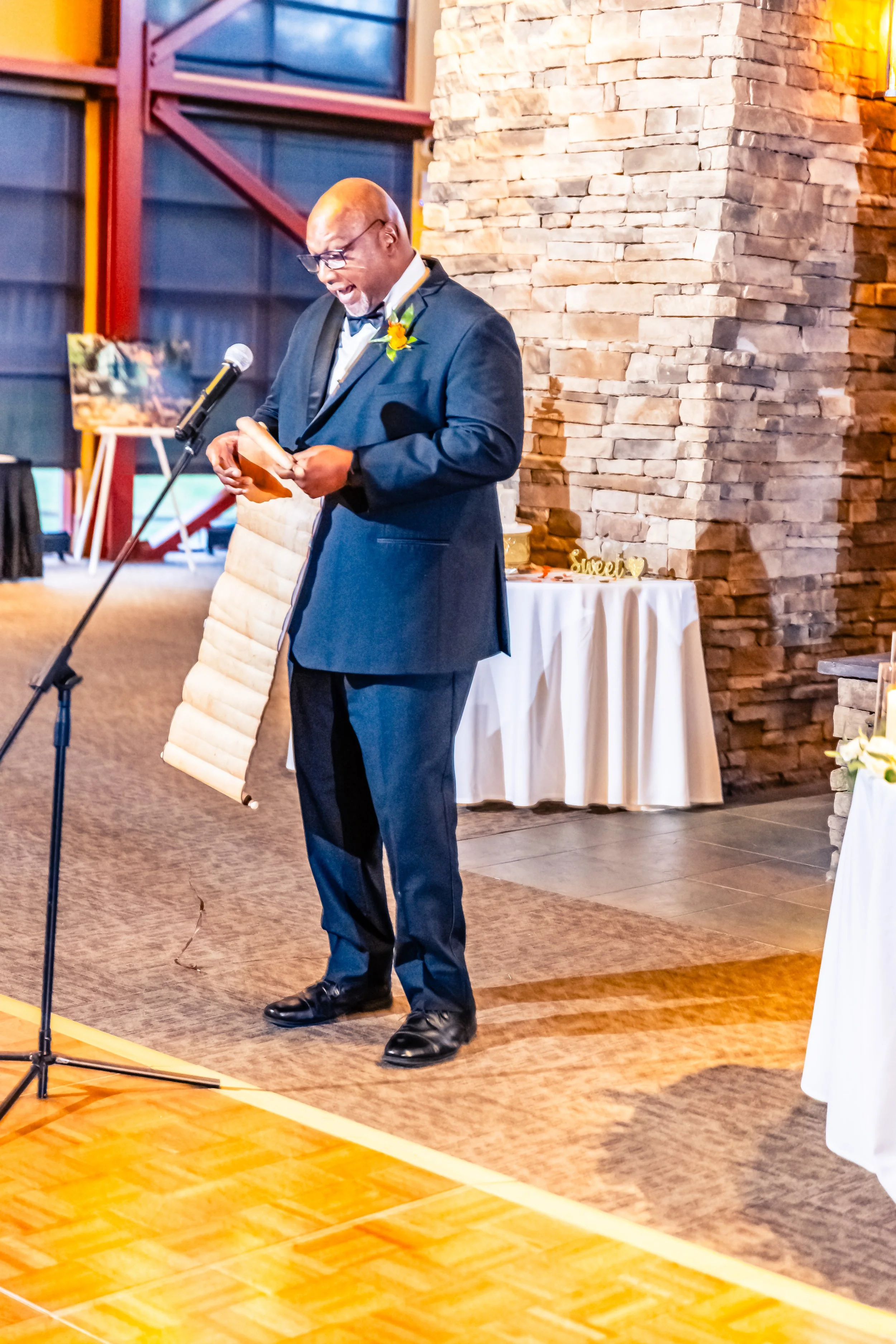 A man in a dark suit with a boutonniere is speaking or reading from a paper at a microphone during a formal event in a venue with a brick wall and tables with white tablecloths.