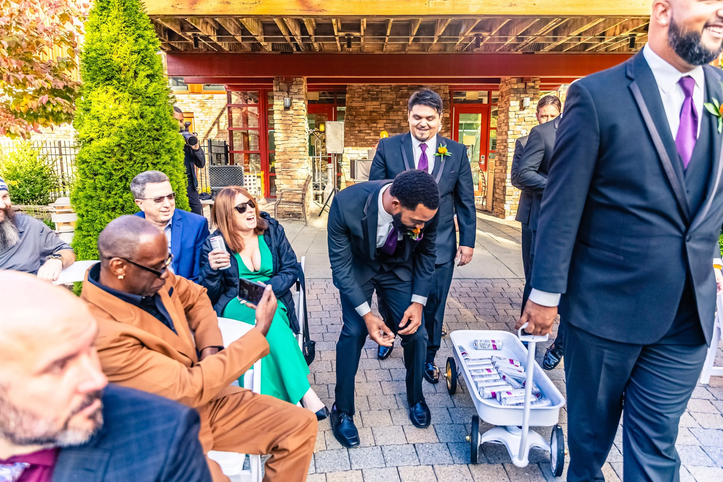 Groom in a suit bending over to pick up objects from a white cart, surrounded by men and women at an outdoor wedding celebration, with some guests sitting and others standing, in front of a building with brick and stone accents.