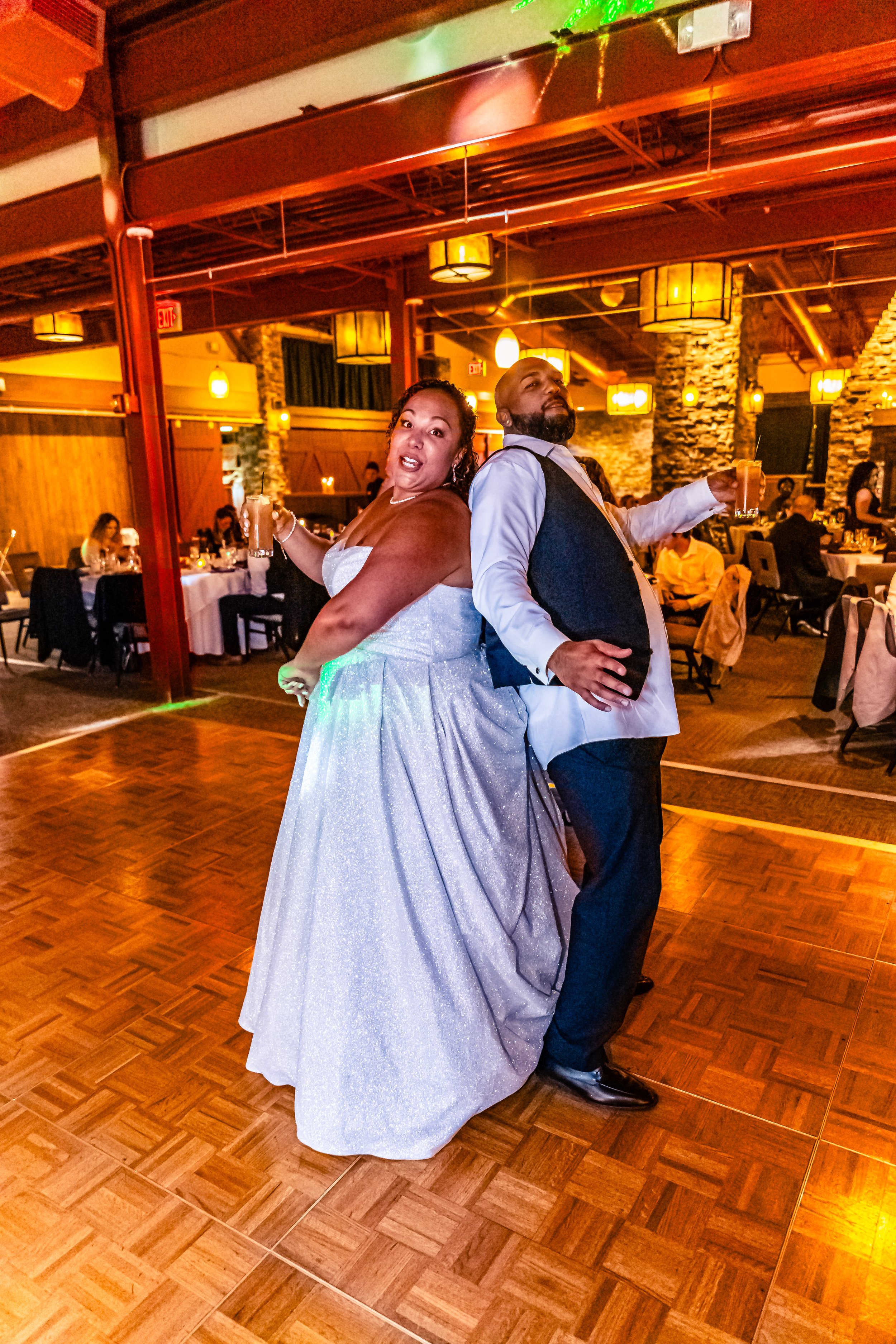 A man and woman dancing at a wedding reception, both holding drinks, with the woman in a white gown and the man in formal attire, in a warmly lit venue with tables and guests in the background.