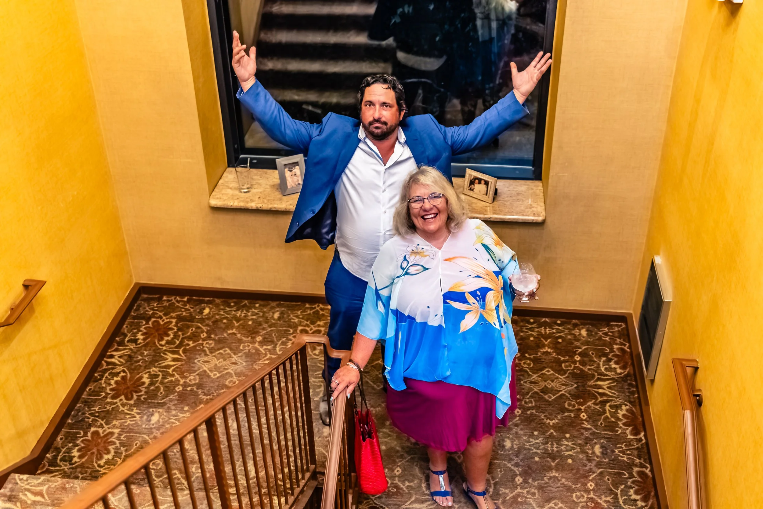 A man in a blue suit with arms raised standing behind a woman in a floral top holding a drink, in an indoor setting with yellow walls and a patterned carpet.