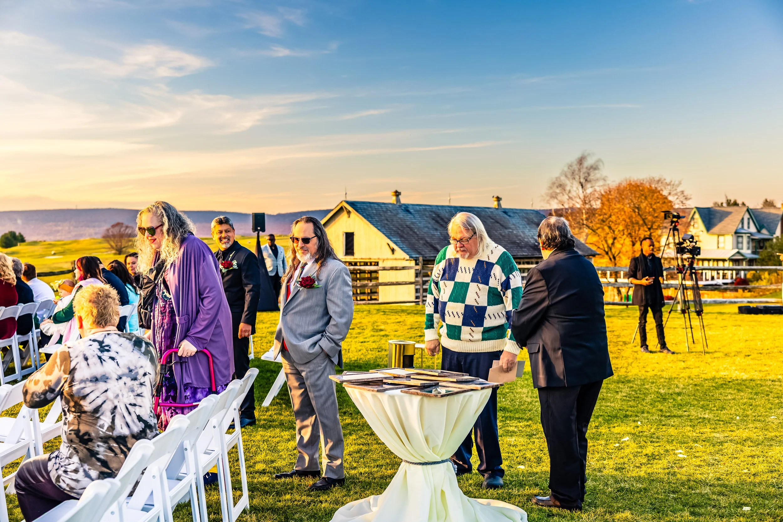 People attending an outdoor ceremony during sunset, some standing in line, others seated, with cameras and a small table with framed photos in the foreground. There are houses and trees in the background.