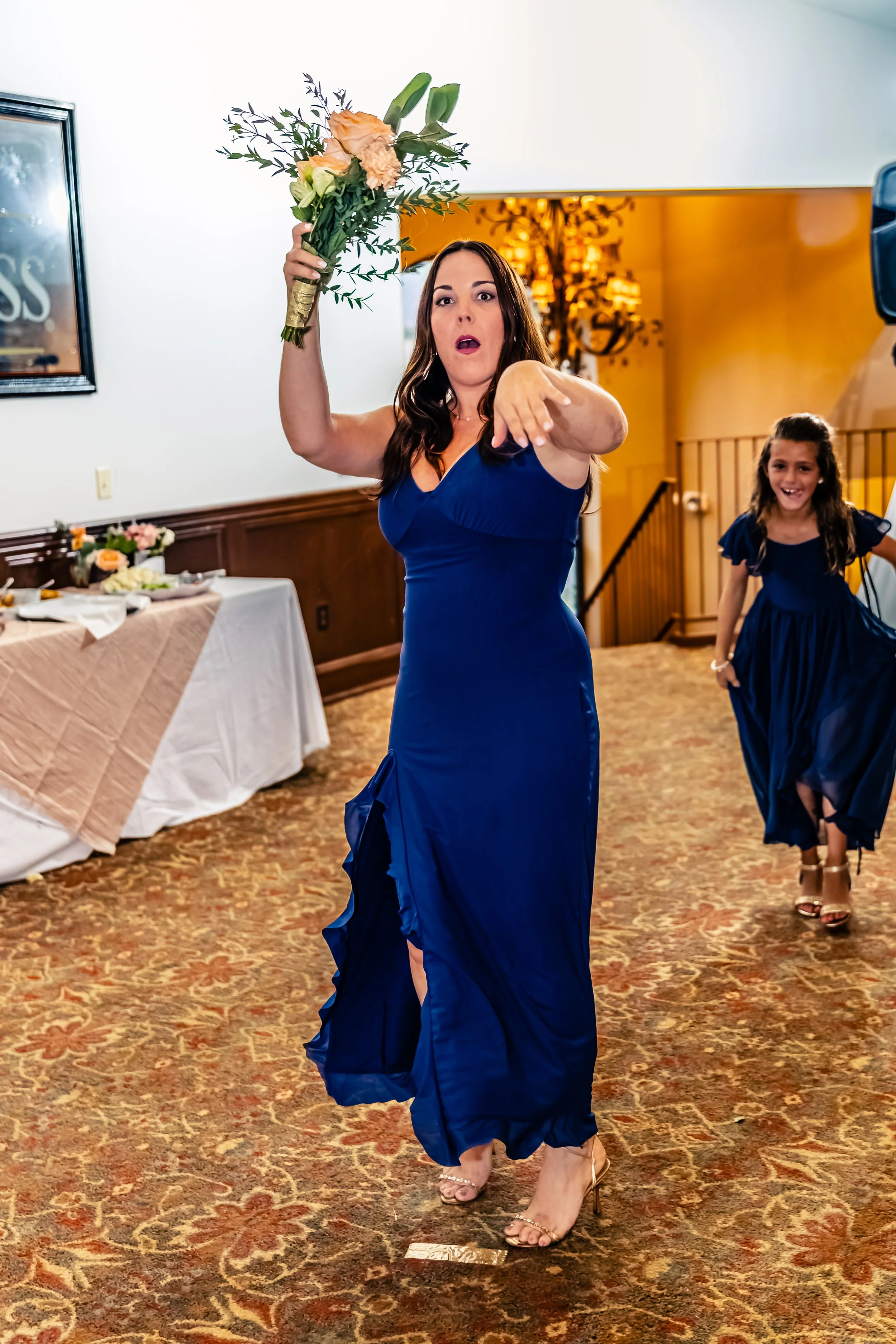 Woman in a blue dress holding a bouquet of flowers, pointing, with a young girl in a matching blue dress behind her, inside a decorated event hall.
