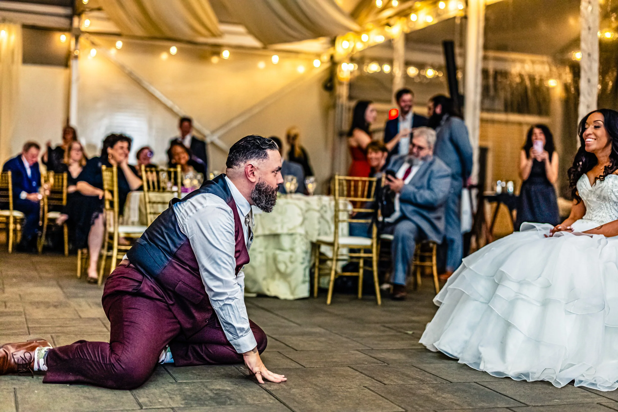 Man kneeling on the floor during wedding reception, facing bride in white gown who is sitting on a chair, with guests seated and standing in the background under a tent with string lights.