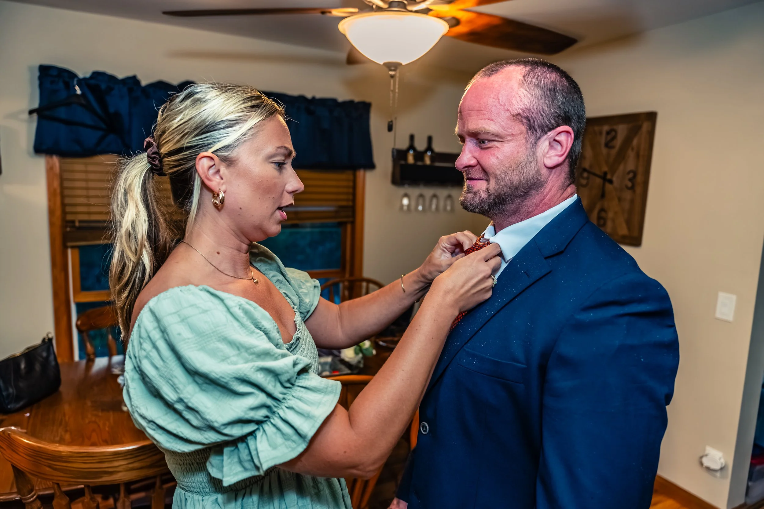 A woman helps a man adjust his tie indoors, with a wooden dining table and chairs, a window with blue curtains, and a wall clock in the background.