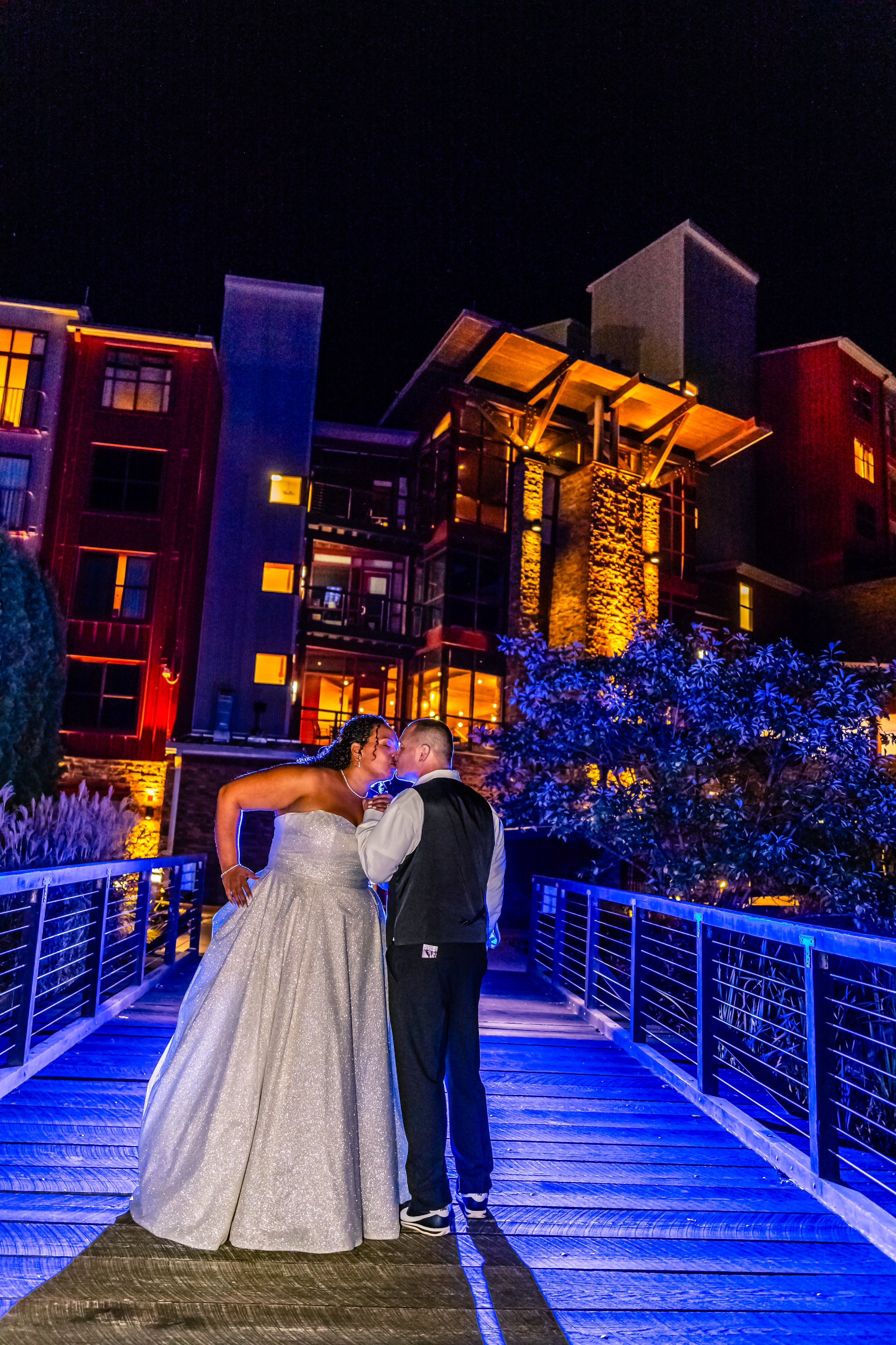 A bride and groom sharing a kiss on a bridge at night, with modern buildings lit up behind them.