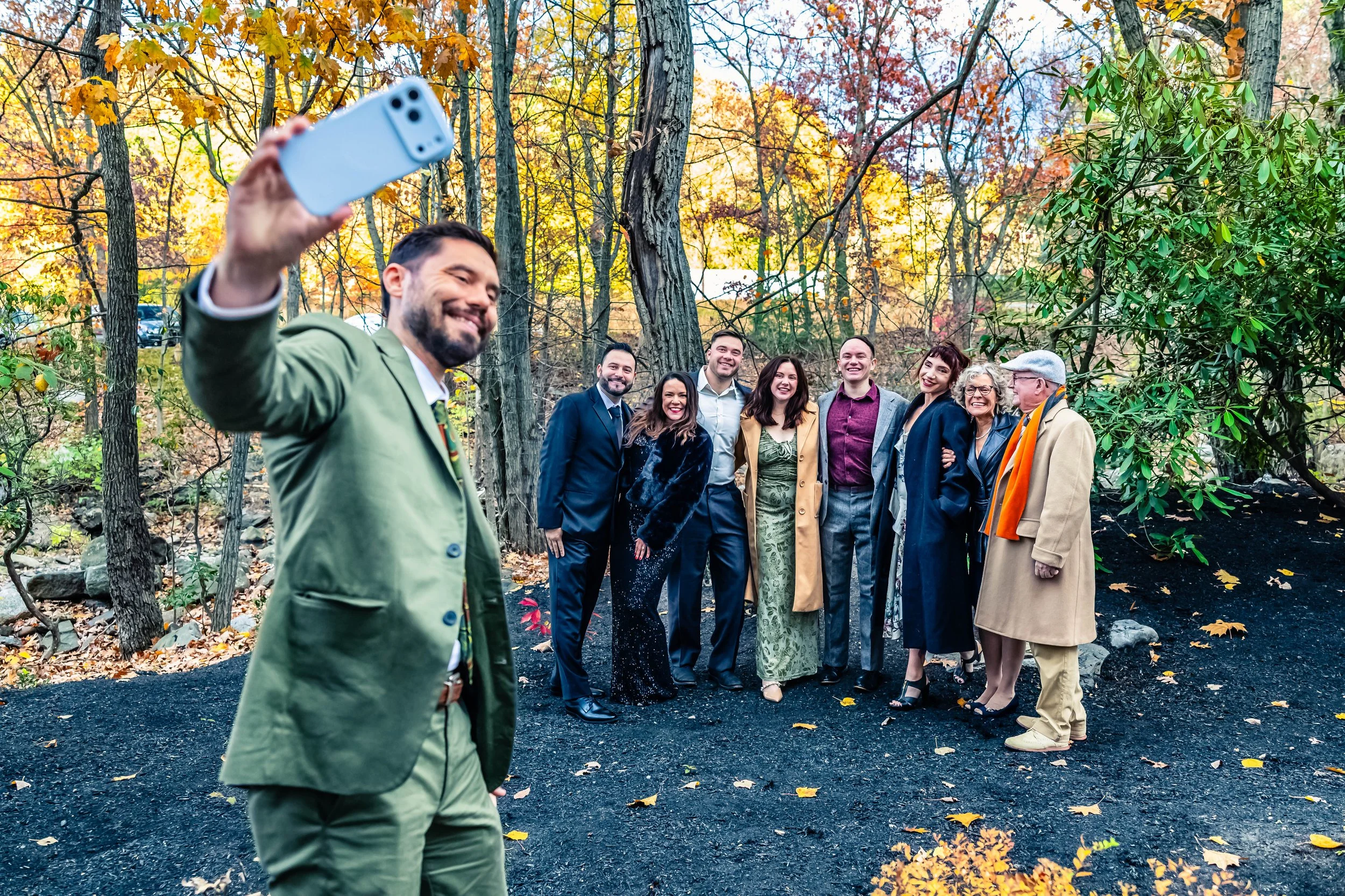 A man wearing a green suit is taking a selfie with a group of eight people in a park during autumn. The group is dressed in formal and semi-formal attire and stands on black gravel among leafless trees and colorful fall foliage.