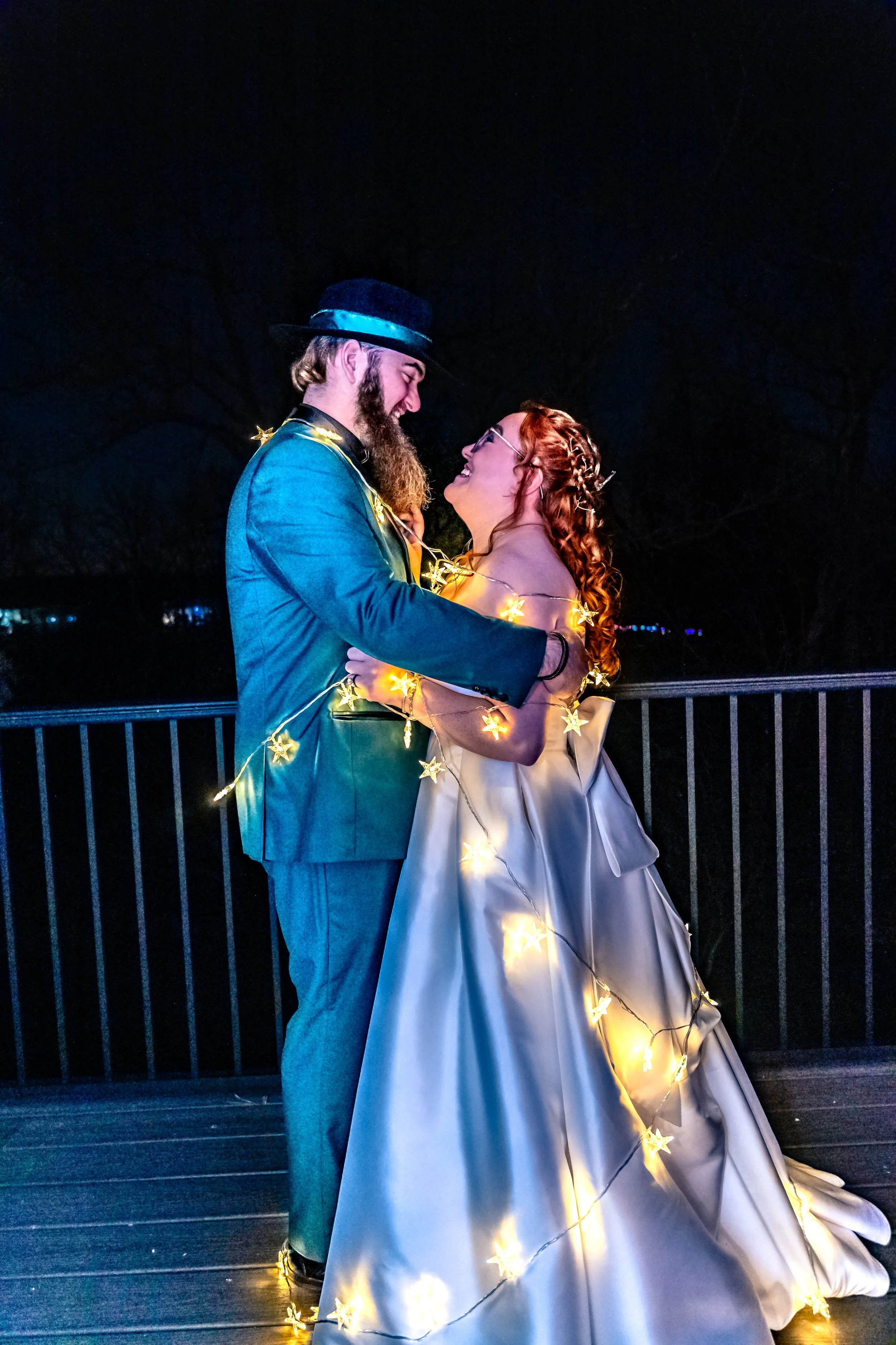 A couple dressed in formal attire, embracing at night, decorated with string lights shaped like stars, standing on a balcony with a dark sky and trees in the background.