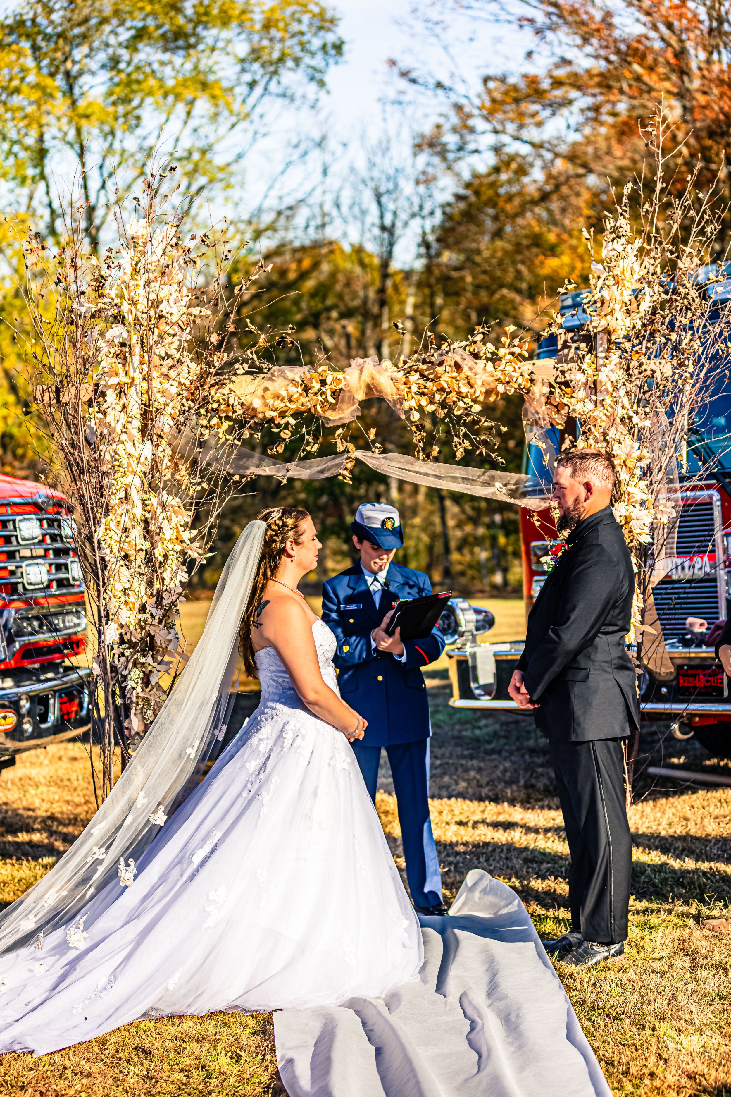 A wedding ceremony taking place outdoors under a decorative floral arch with a bride and groom exchanging vows, an officiant in a military uniform officiant standing between them, and fire trucks parked in the background.