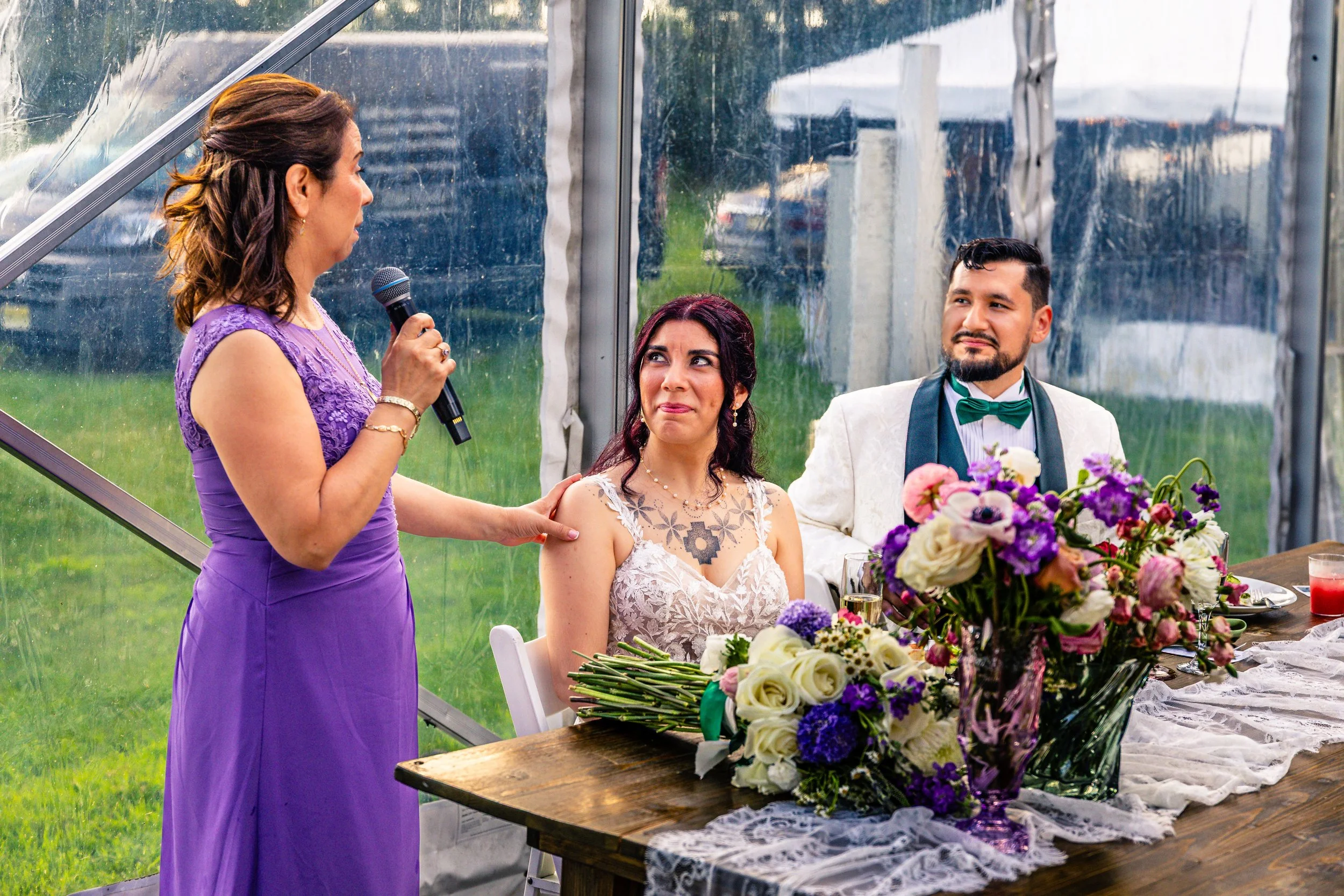 A woman in a purple dress giving a speech at a wedding reception, with a bride and groom sitting at a table decorated with flowers and cups, inside a transparent tent.