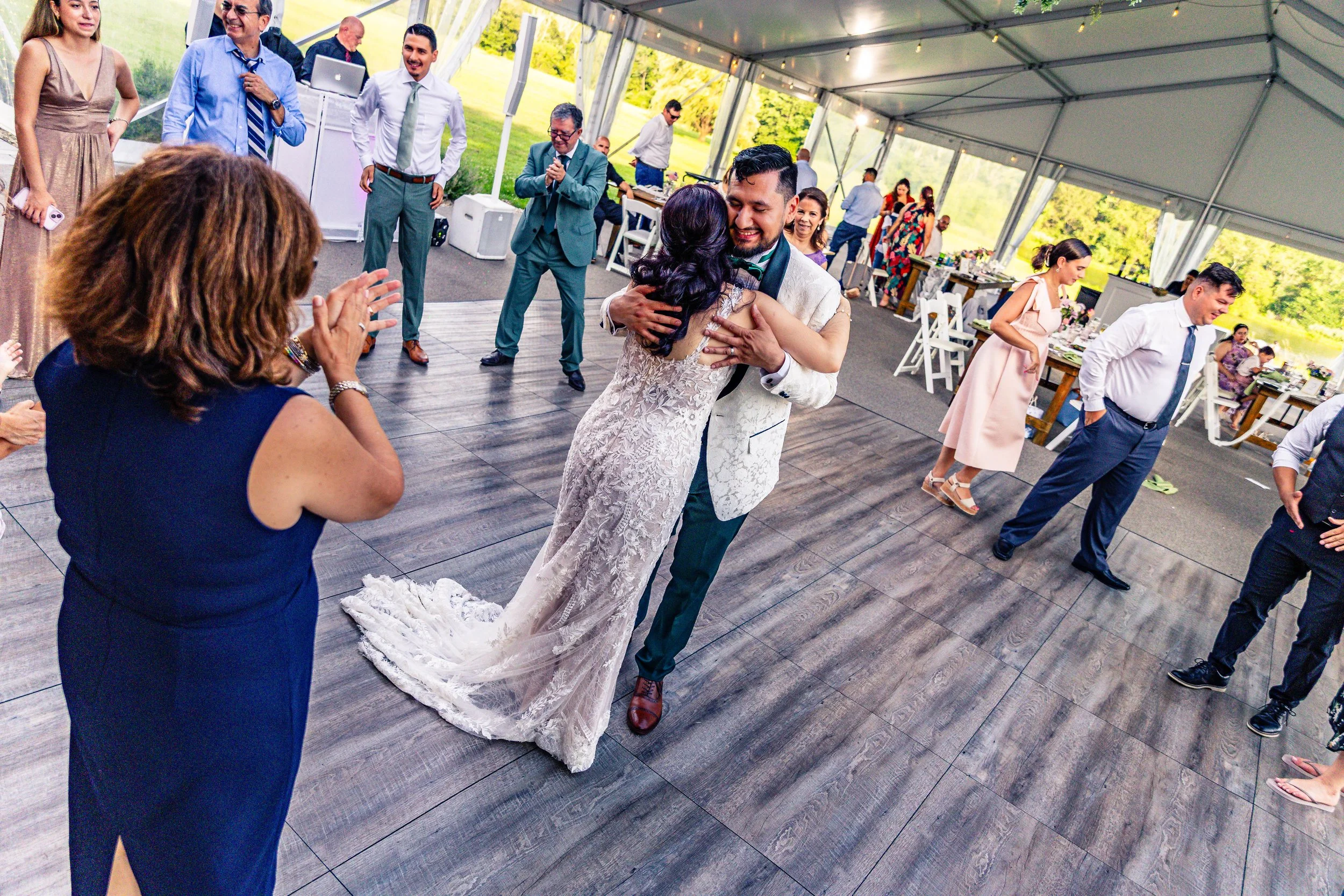 A wedding reception with guests dancing and socializing inside a large tent with a wooden dance floor and outdoor views. The bride and groom are dancing-center, surrounded by friends and family.