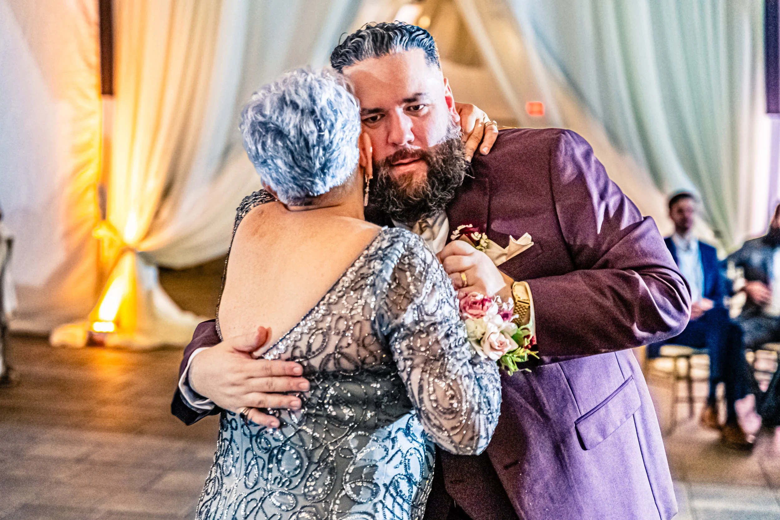 A man and a woman share a dance, embracing each other closely at a festive event, with the woman wearing a glittering dress and the man in a purple suit, surrounded by colorful lighting.
