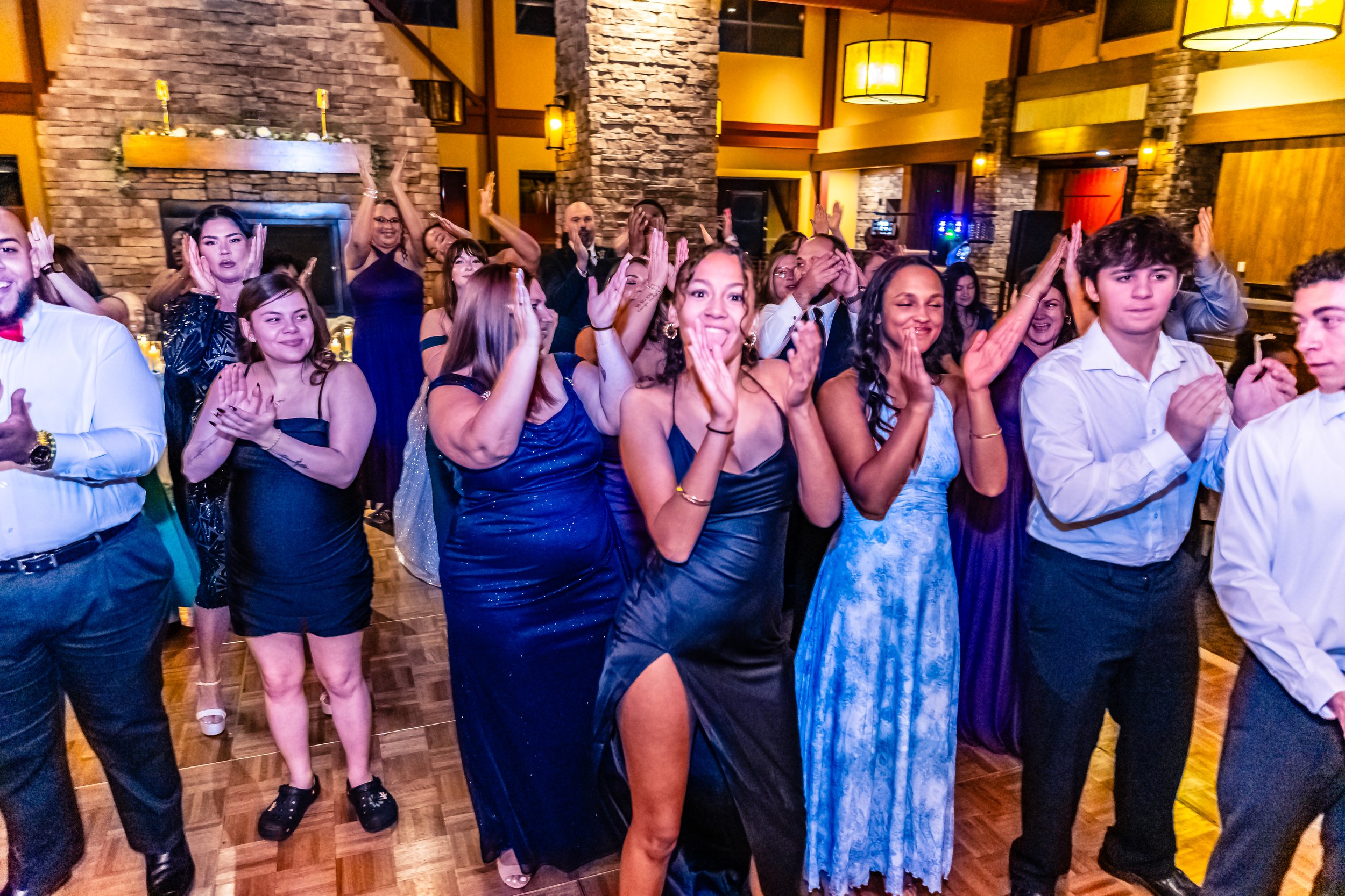 Group of people at a celebration or party, clapping and smiling in a decorated indoor venue with warm lighting and a stone fireplace in the background.