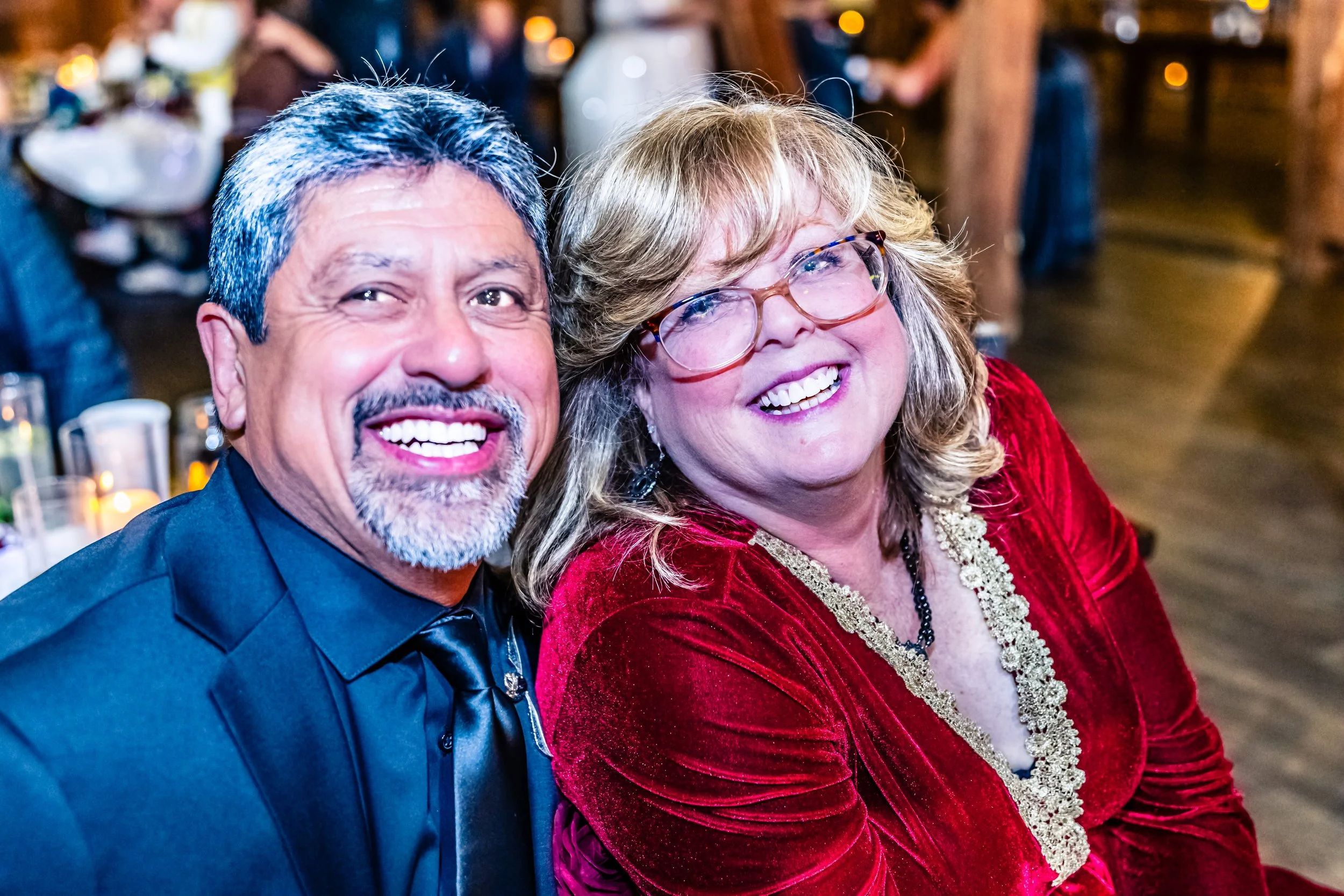 Two smiling people, a man with gray hair and a beard in a black tuxedo, and a woman with blonde hair and glasses in a red dress, at a social event or celebration.